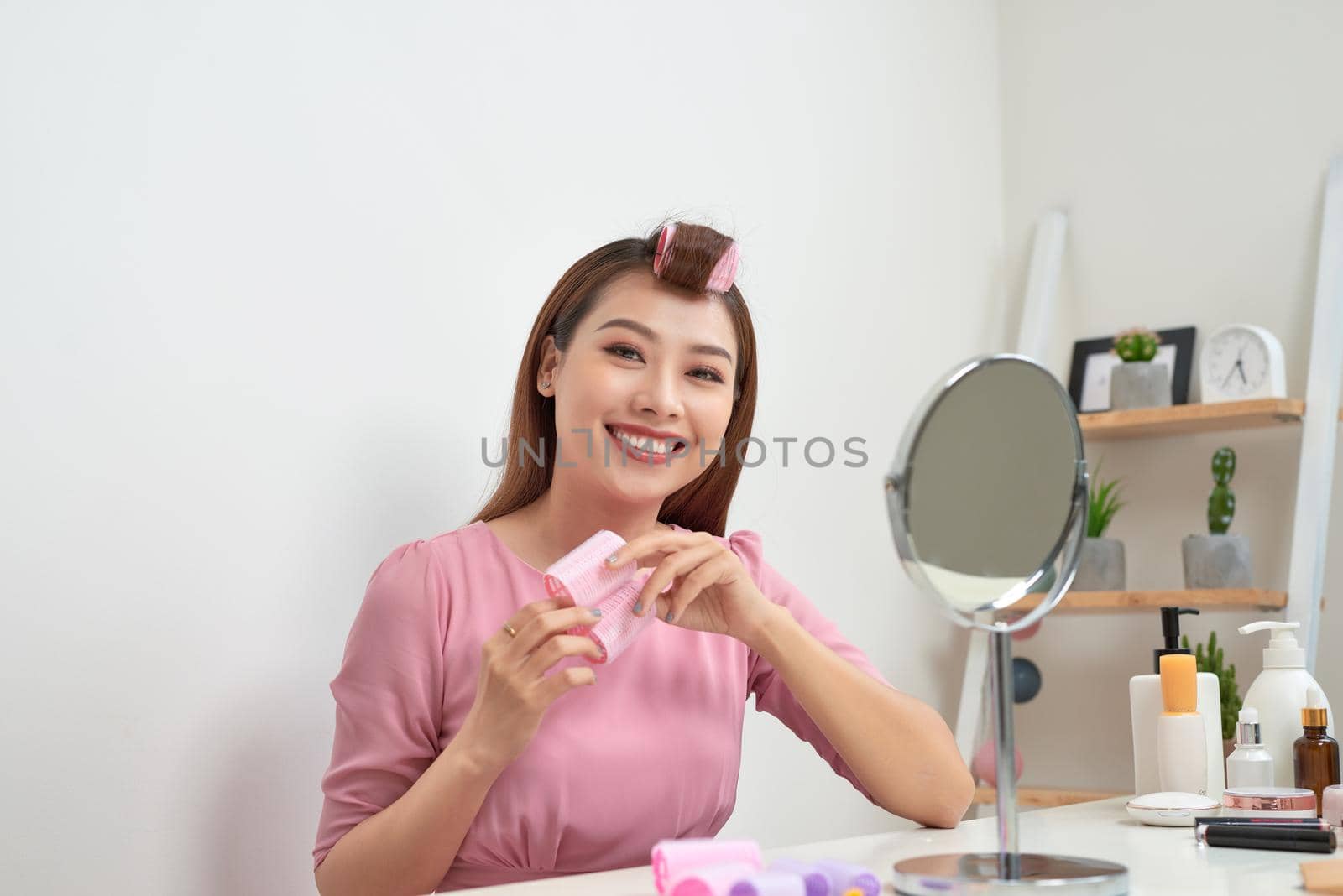 Head and shoulders portrait of beautiful Asian woman wearing hair curlers looking in mirror with wide smile, home interior on background