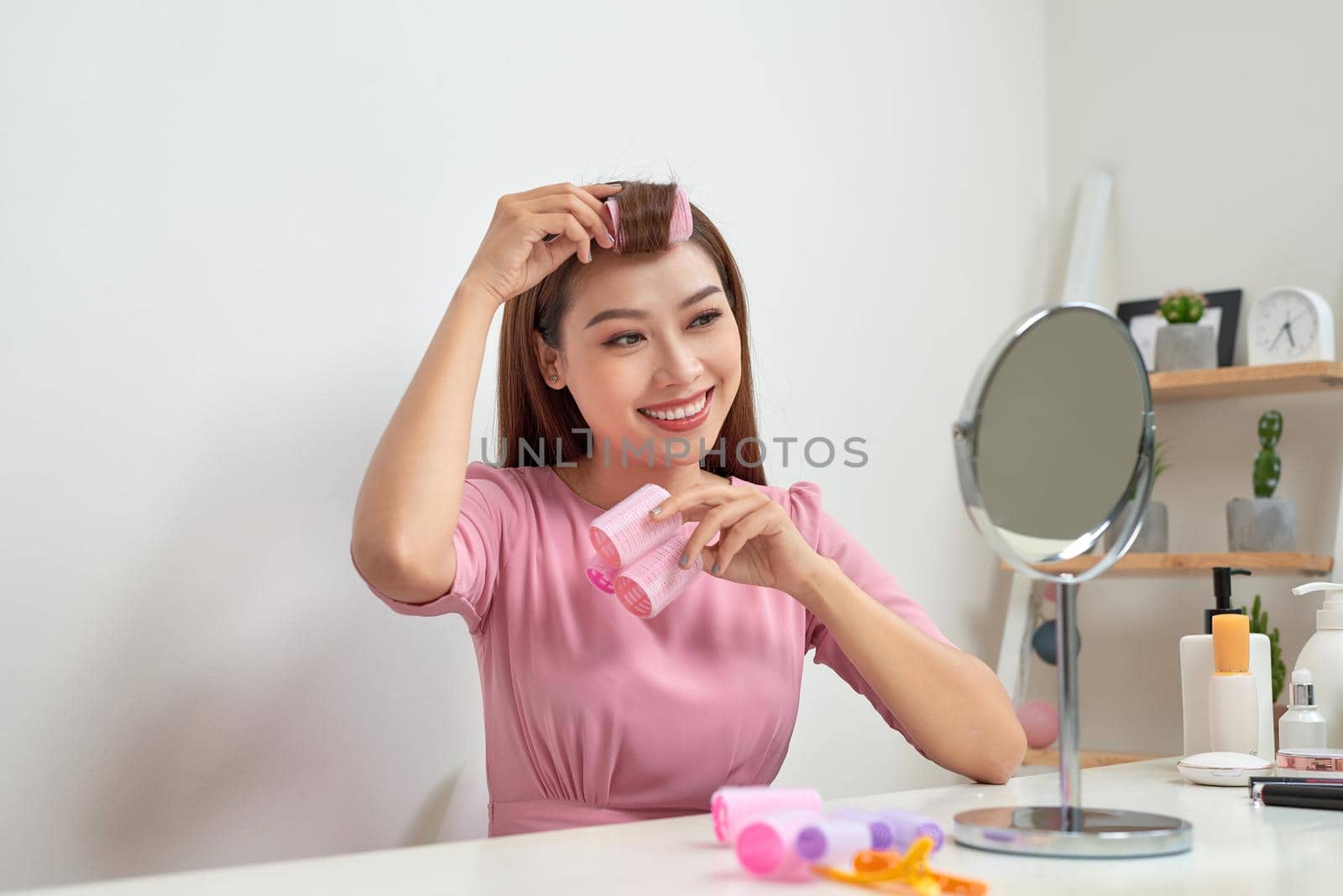 Head and shoulders portrait of beautiful Asian woman wearing hair curlers looking in mirror with wide smile, home interior on background by makidotvn