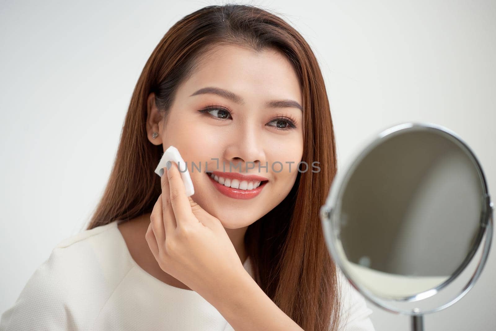 Taking off her make-up. Beautiful cheerful young woman using cotton disk and looking at her reflection in mirror with smile while sitting at the dressing table