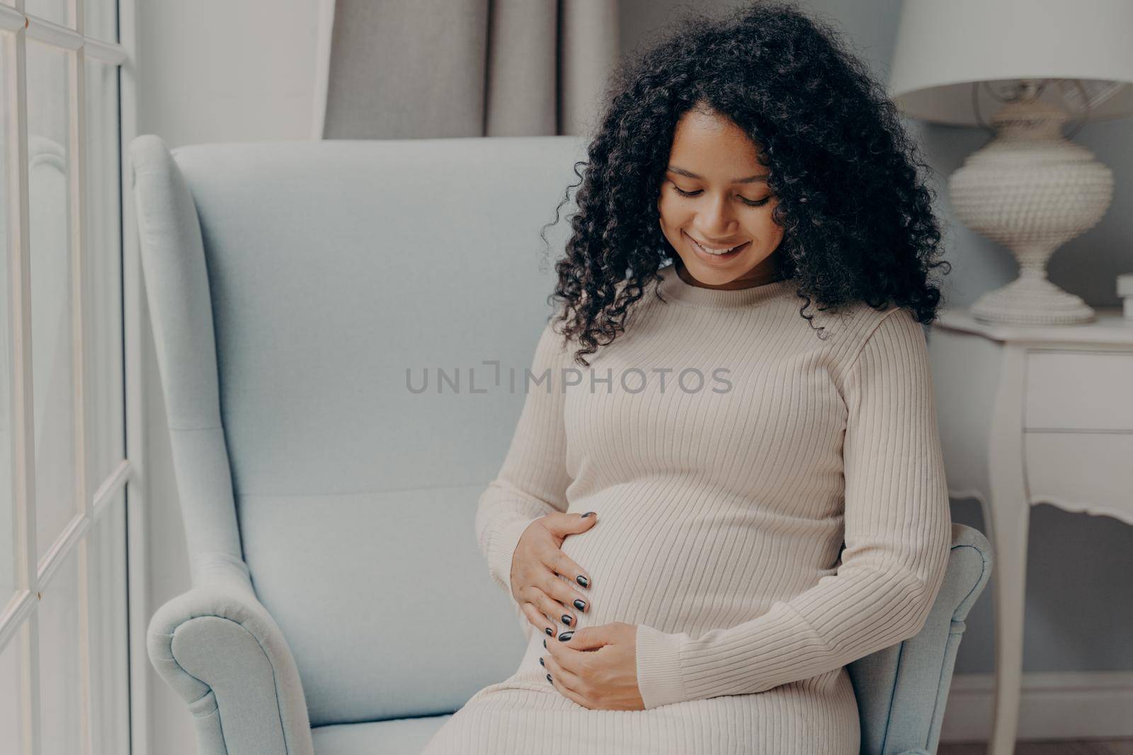 Happy pregnancy. Pretty mixed race woman with curly hair in white casual dress holding her belly in anticipation of future baby, thinking about maternity and how to be caring and loving mother