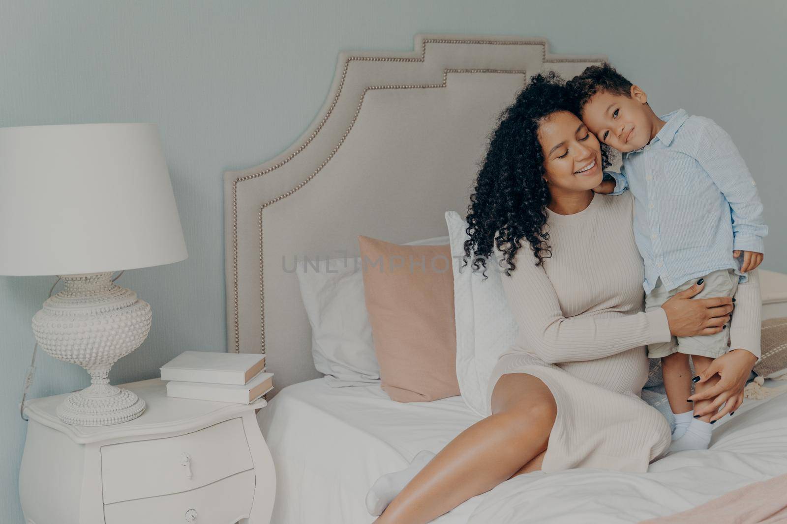 My little boy. Happy pregnant afro american woman in casual white dress with curly hair sitting on bed with her sweet little son and showing strong feelings of love and care. Maternity and pregnancy