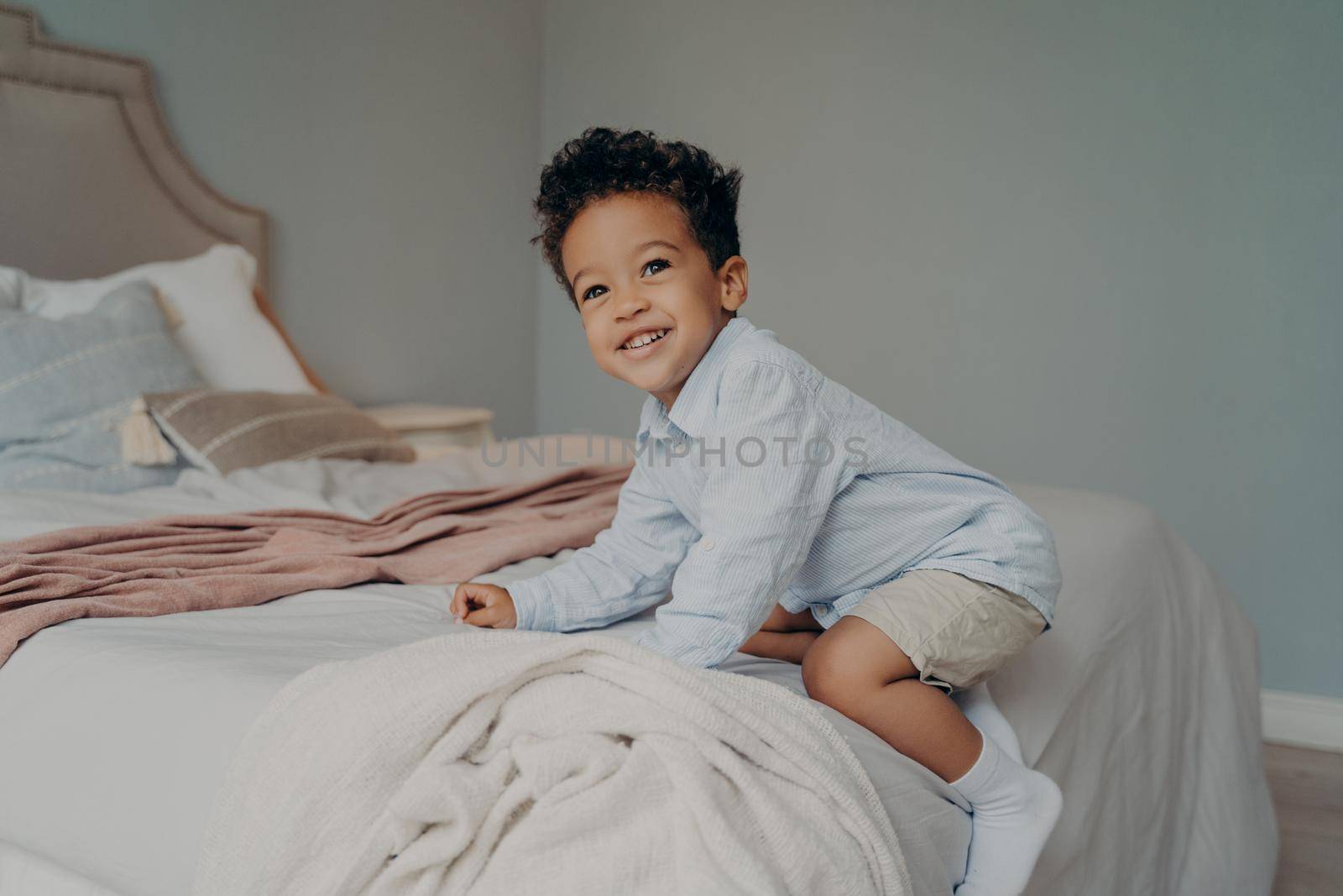 Cute happy african american kid boy playing and having fun on bed after coming home from kindergarten, playing and smiling while spending time in bedroom. Carefree childhood concept