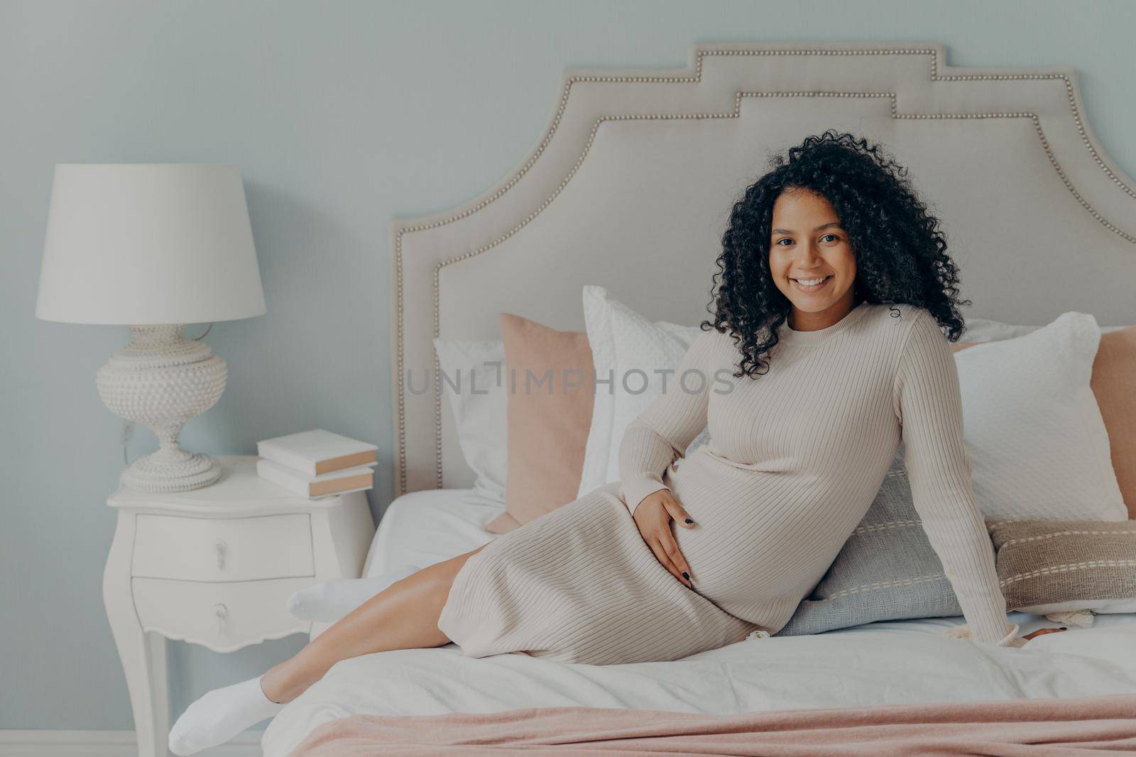 Cheerful happy pregnant mixed race future mom with curly hair relaxing on big comfy bed after long trip to shopping mall, being on last months of pregnancy, holding her belly with one hand and smiling