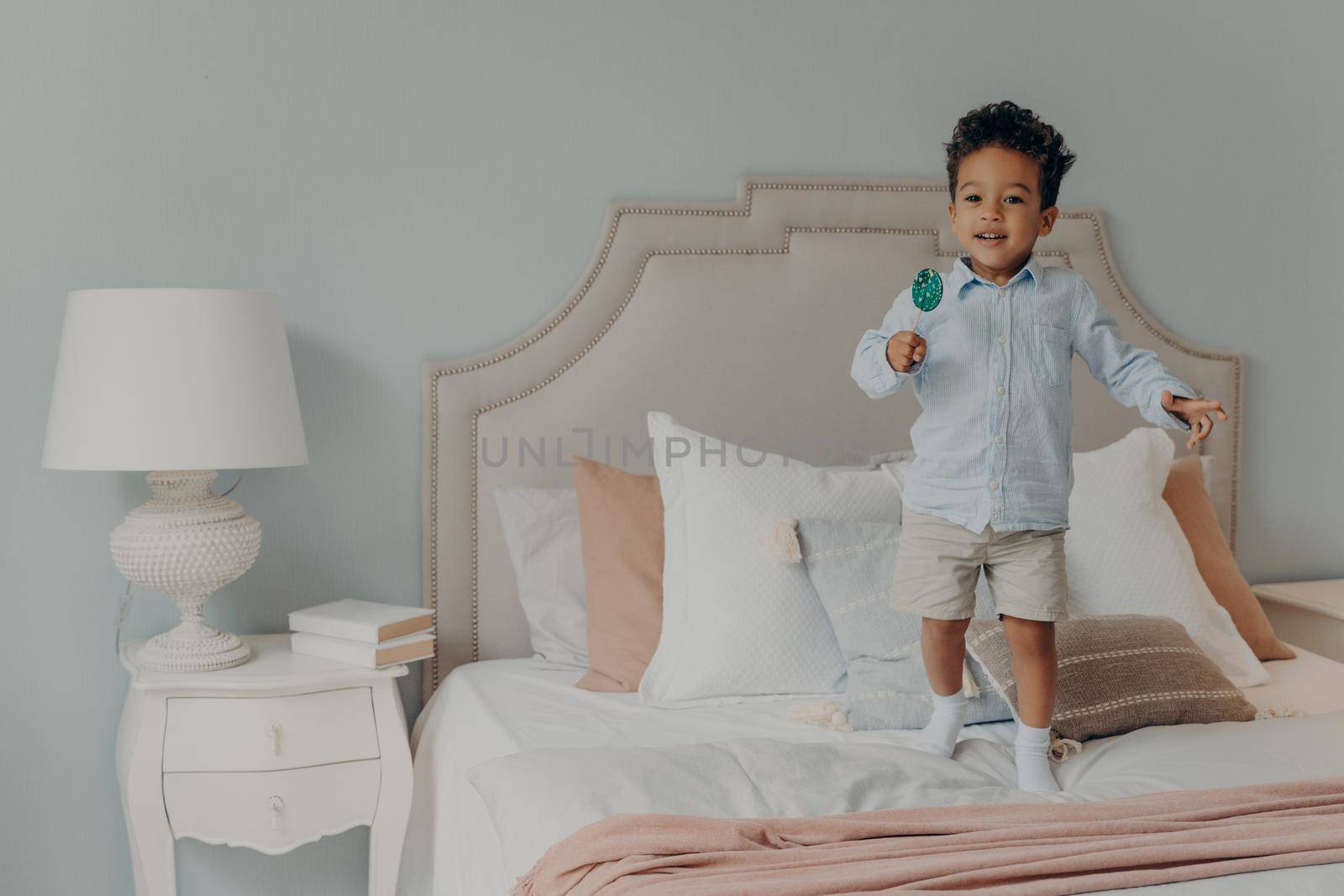 Happy playful afro american child boy with lollipop having lots of fun in bedroom, jumping on bed mattress with variety of pillows and eating candy while mom is not watching. Childhood concept