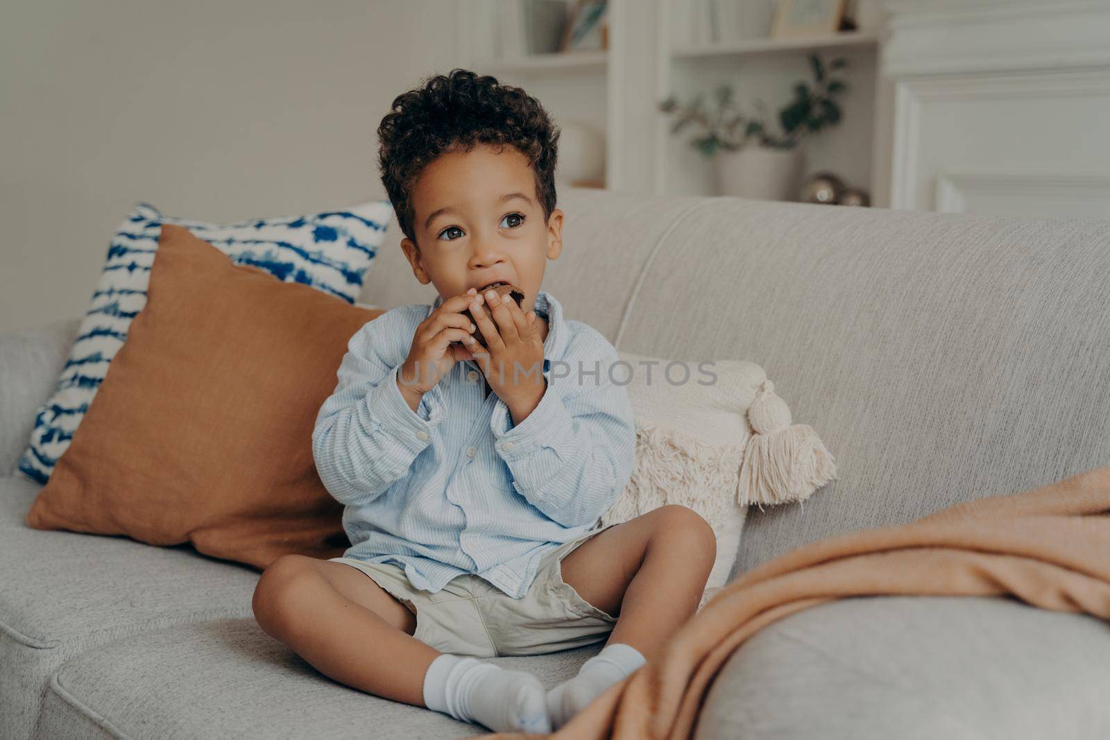 Hungry cute african american boy toddler eating delicious chocolate cupcake that mother brought from local bakery, enjoying the sweet taste while sitting on light sofa in living room at home