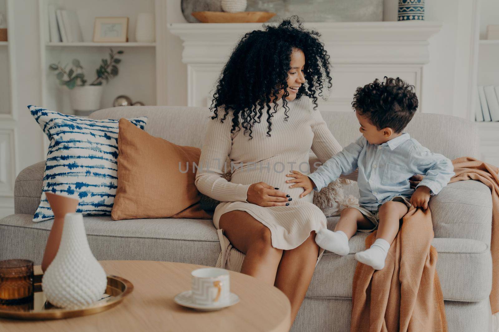 Cute little boy touching belly of his pregnant mom while relaxing together on sofa in living room by vkstock