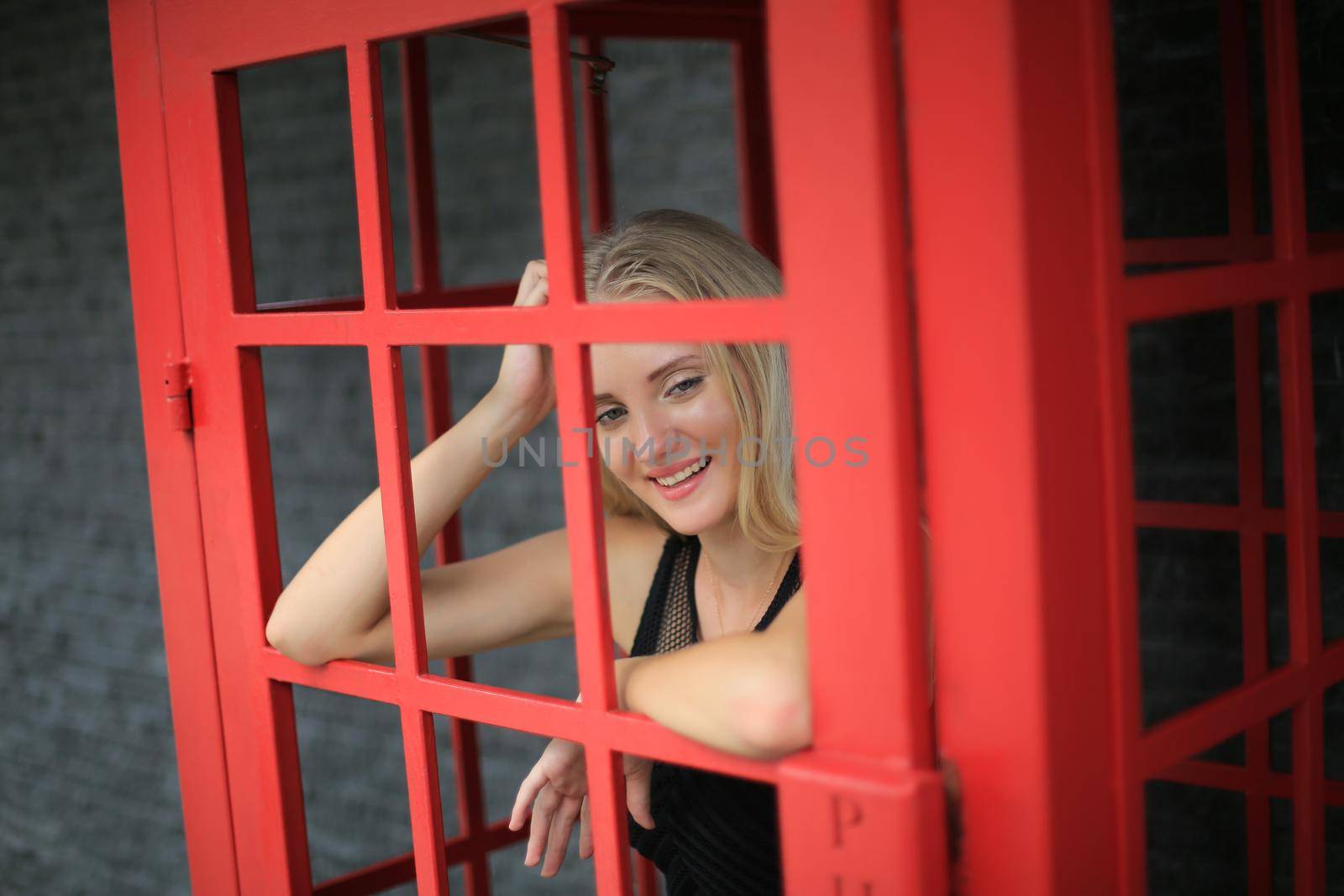 Portrait of Beautiful blonde hair girl on black dress standing in red phone booth against black wall as portrait fashion pose outdoor. by chuanchai