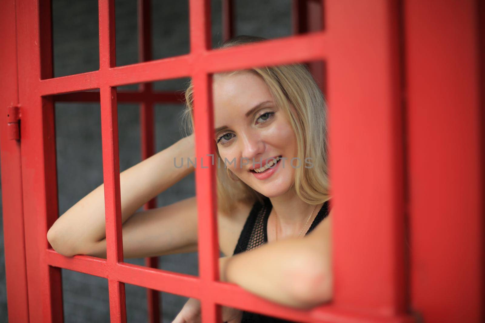 Portrait of Beautiful blonde hair girl on black dress standing in red phone booth against black wall as portrait fashion pose outdoor. by chuanchai