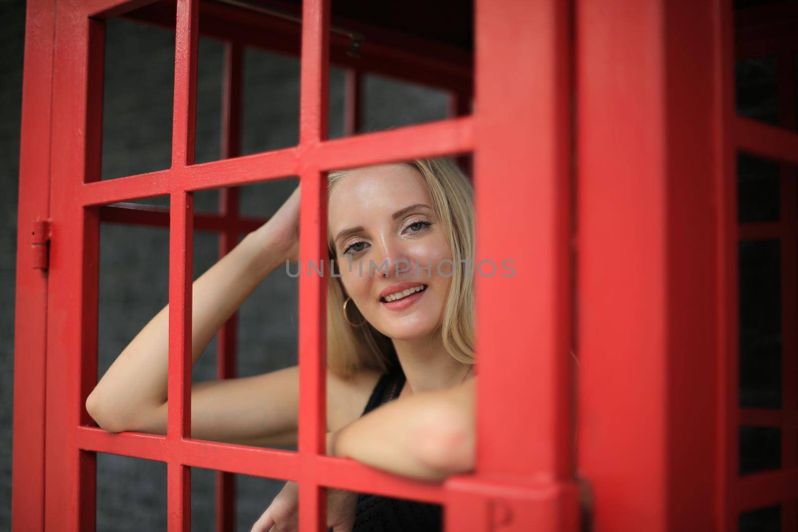 Portrait of Beautiful blonde hair girl on black dress standing in red phone booth against black wall as portrait fashion pose outdoor. by chuanchai