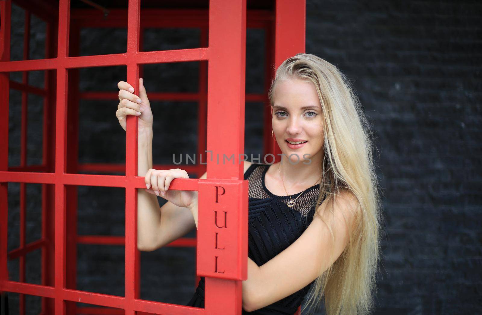 Portrait of Beautiful blonde hair girl on black dress standing in red phone booth against black wall as portrait fashion pose outdoor.