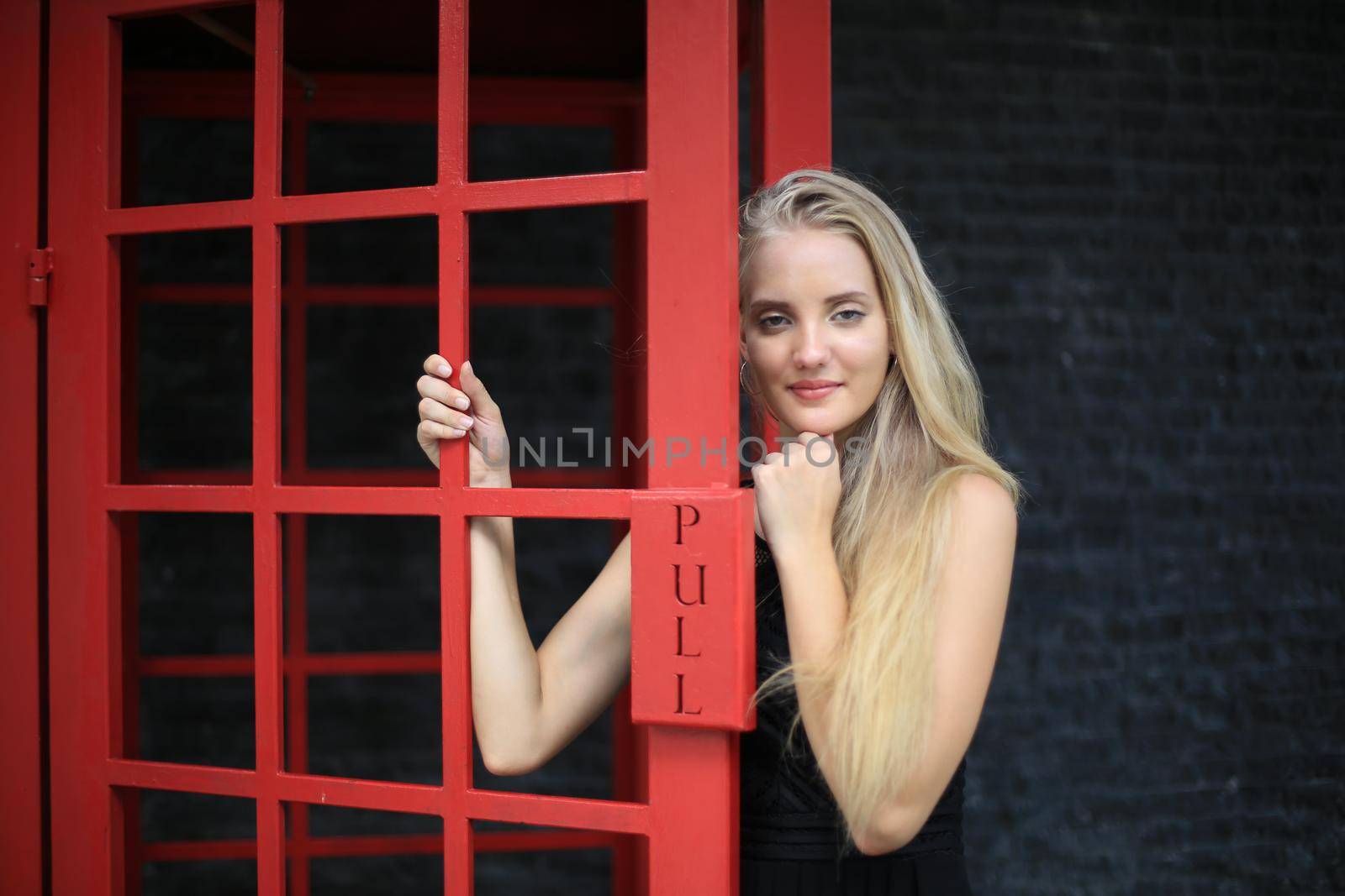 Portrait of Beautiful blonde hair girl on black dress standing in red phone booth against black wall as portrait fashion pose outdoor. by chuanchai