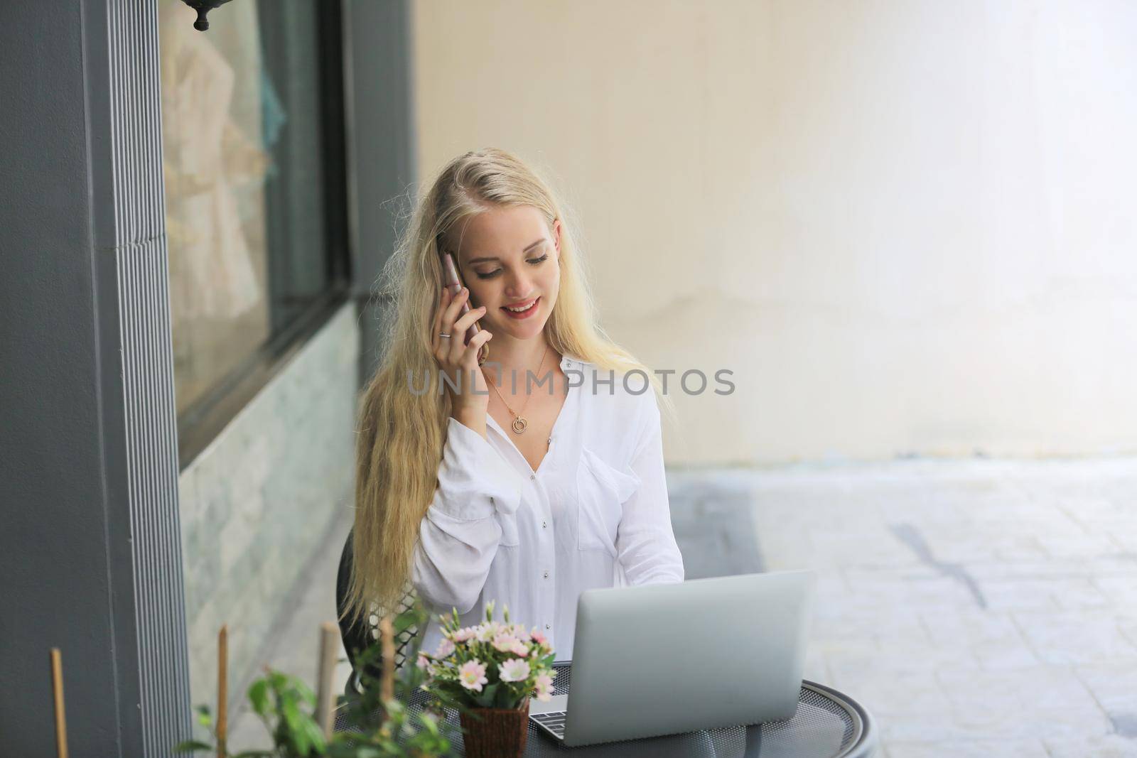 Beautiful blonde hair girl speaking on mobile phone with laptop on table outside coffee shop.