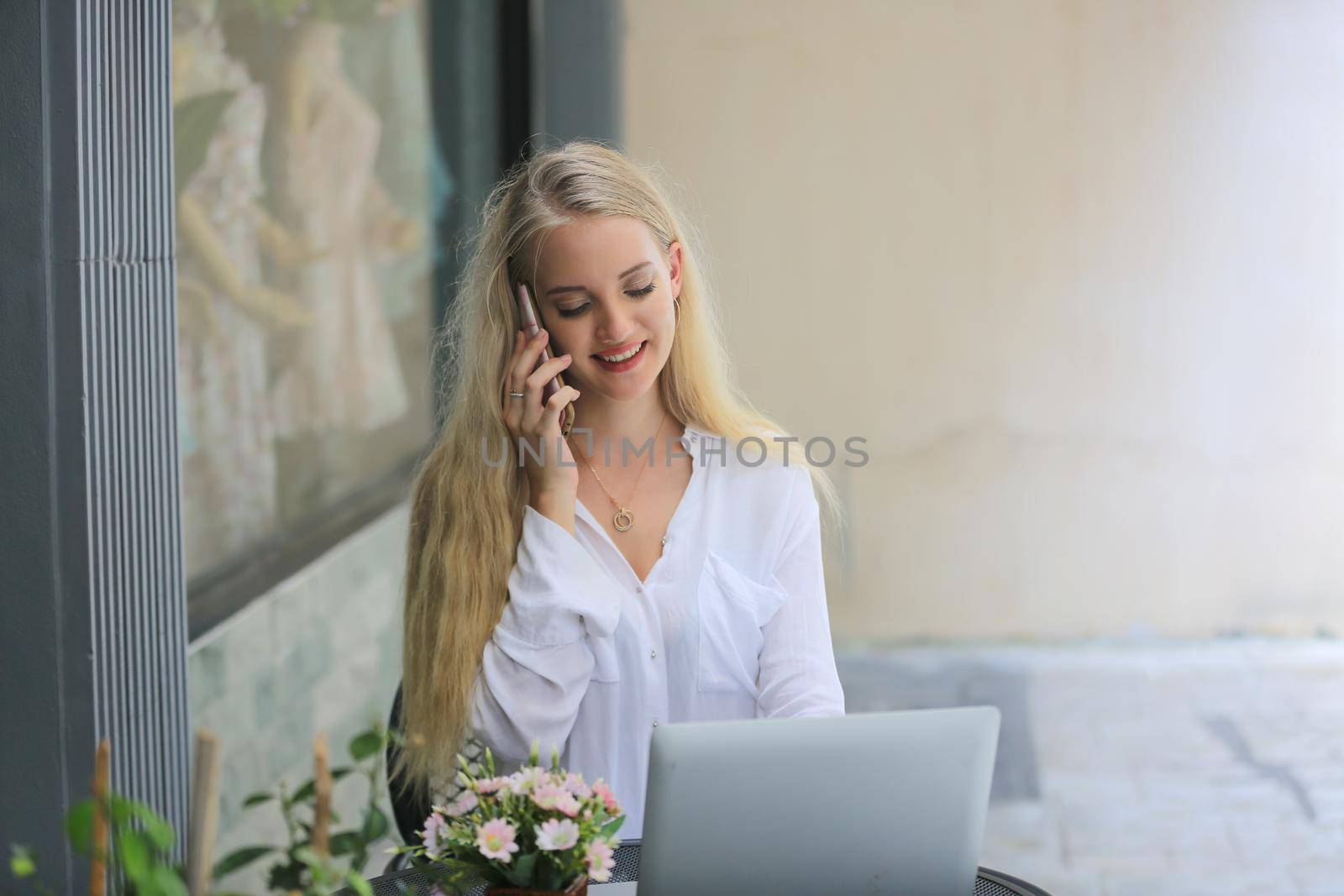 Beautiful blonde hair girl speaking on mobile phone with laptop on table outside coffee shop. by chuanchai