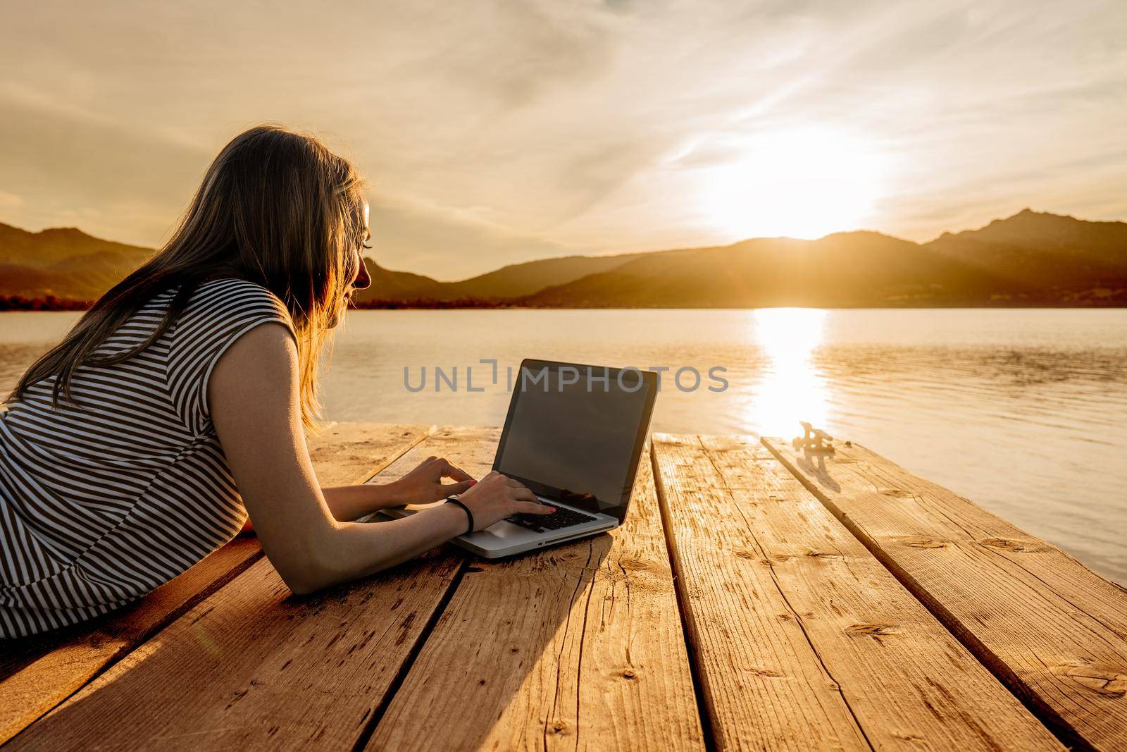 Young woman enjoying her creativeness writing book on a pier at sunset. New job opportunity at modern times to work everywhere using laptop and wifi internet connection technology. Girl studying