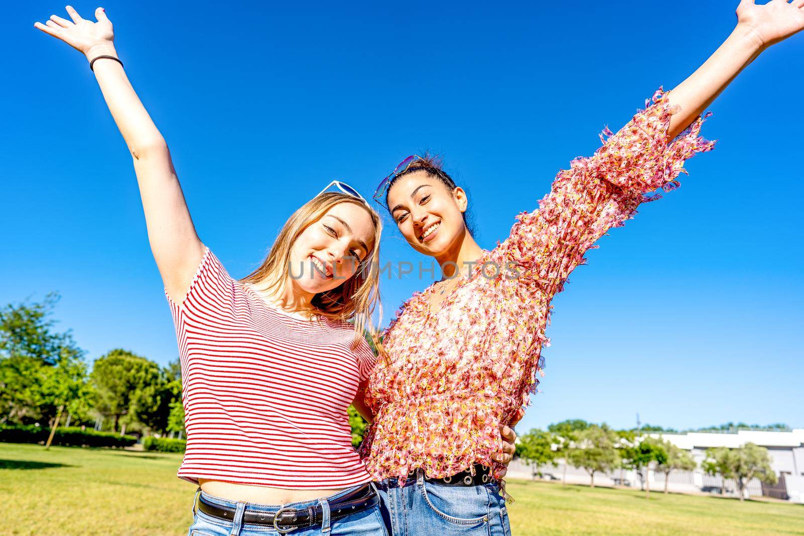 Two happy girls embracing each other with open up arms smiling looking at camera in a city park field in sunny day with blue sky. Mixed young women couple enjoying nature celebrating their diversity by robbyfontanesi