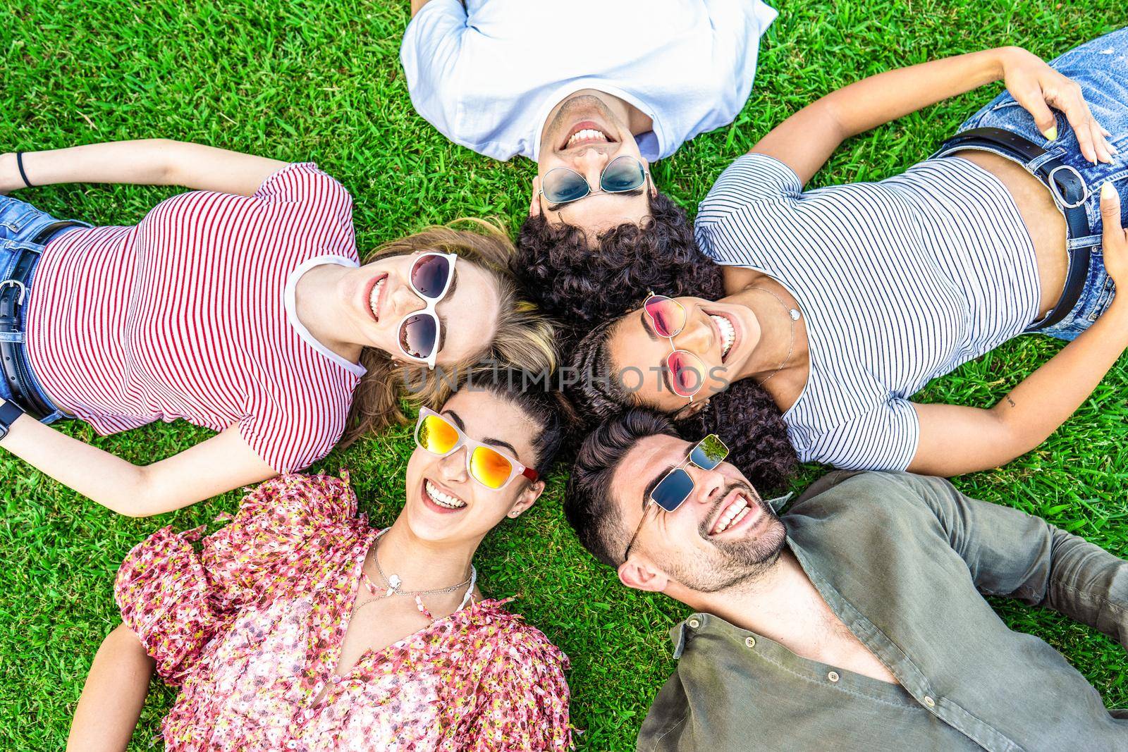 Group of friends lying on park meadow wearing colored trendy sunglasses looking at camera laughing. Happy diverse international people enjoying friendship in nature resting in circle. Mixed race union