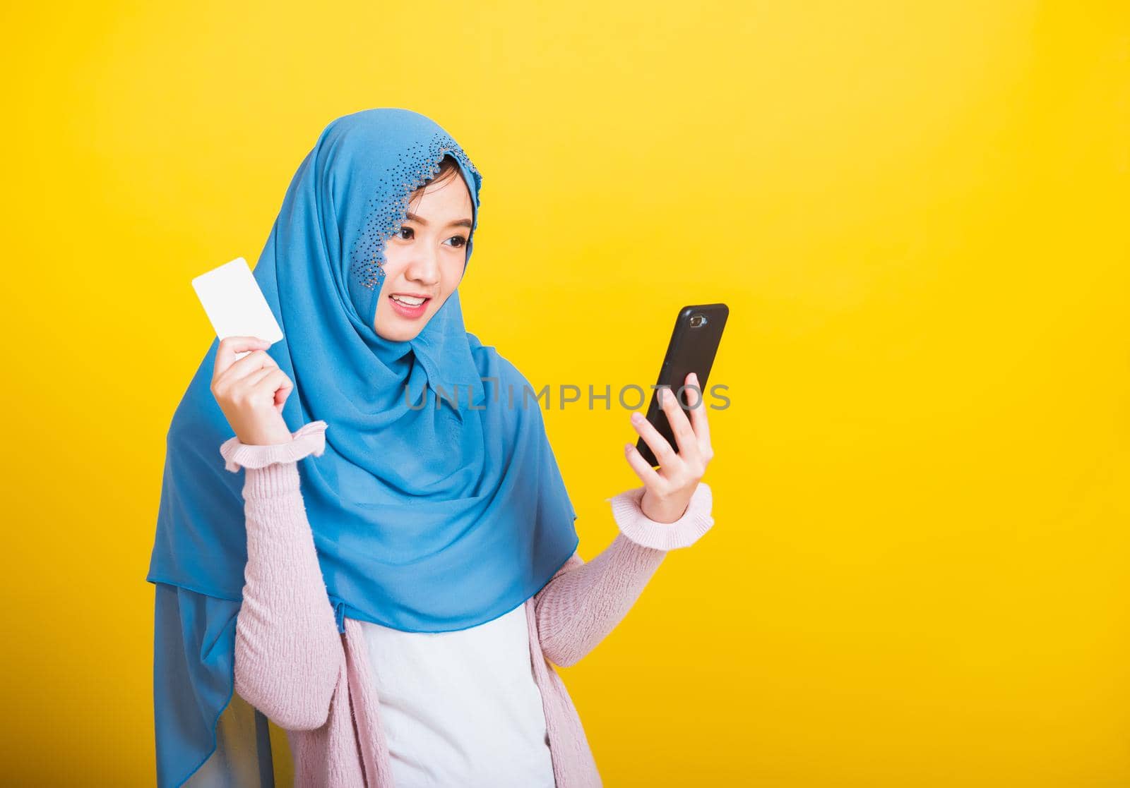 Asian Muslim Arab, Portrait of happy beautiful young woman Islam religious wear veil hijab funny smile she using mobile phone and credit bank card, studio shot isolated on yellow background