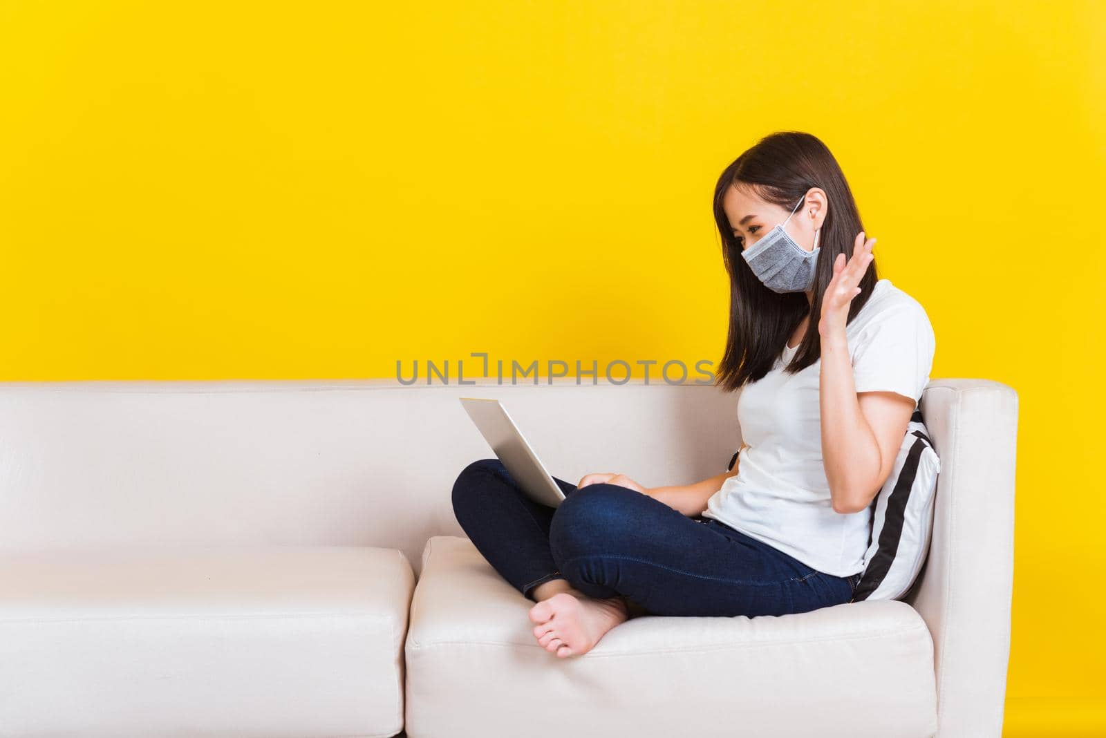 Portrait Asian of beautiful young woman sitting on sofa wearing medical face mask protective she work from home with laptop computer during Coronavirus studio shot isolated on yellow background