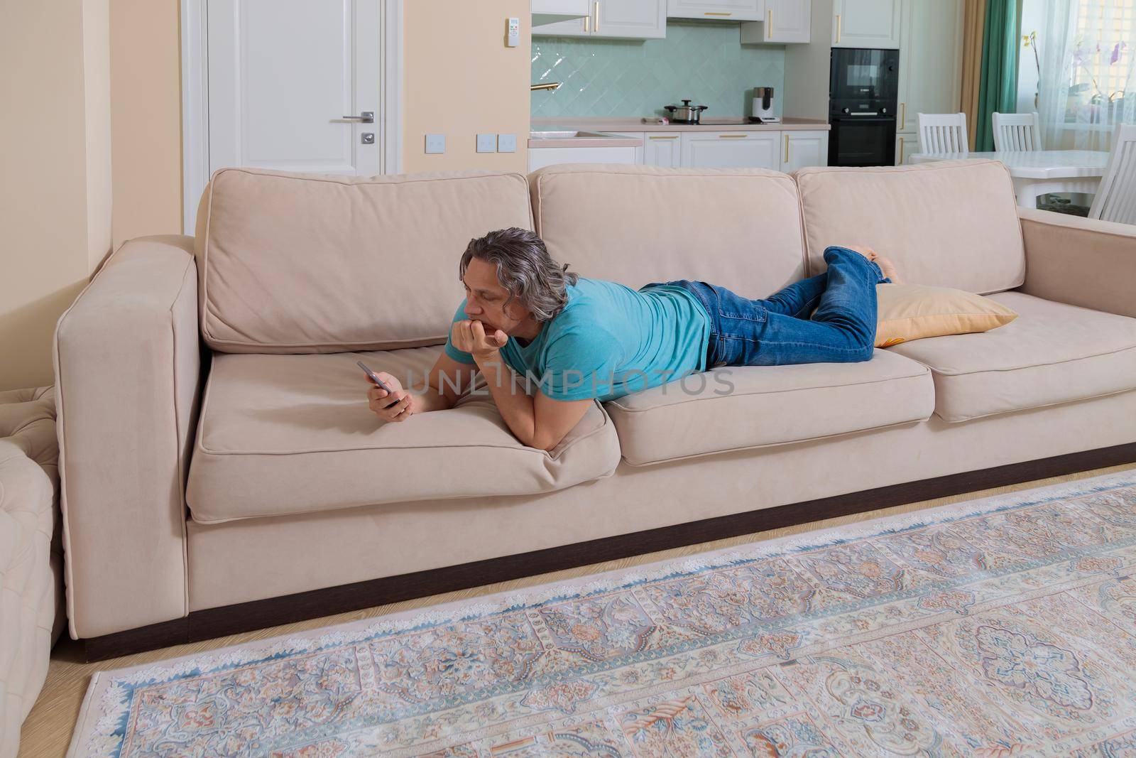 An adult man browses social networks in a mobile phone while lying on a sofa in an apartment.