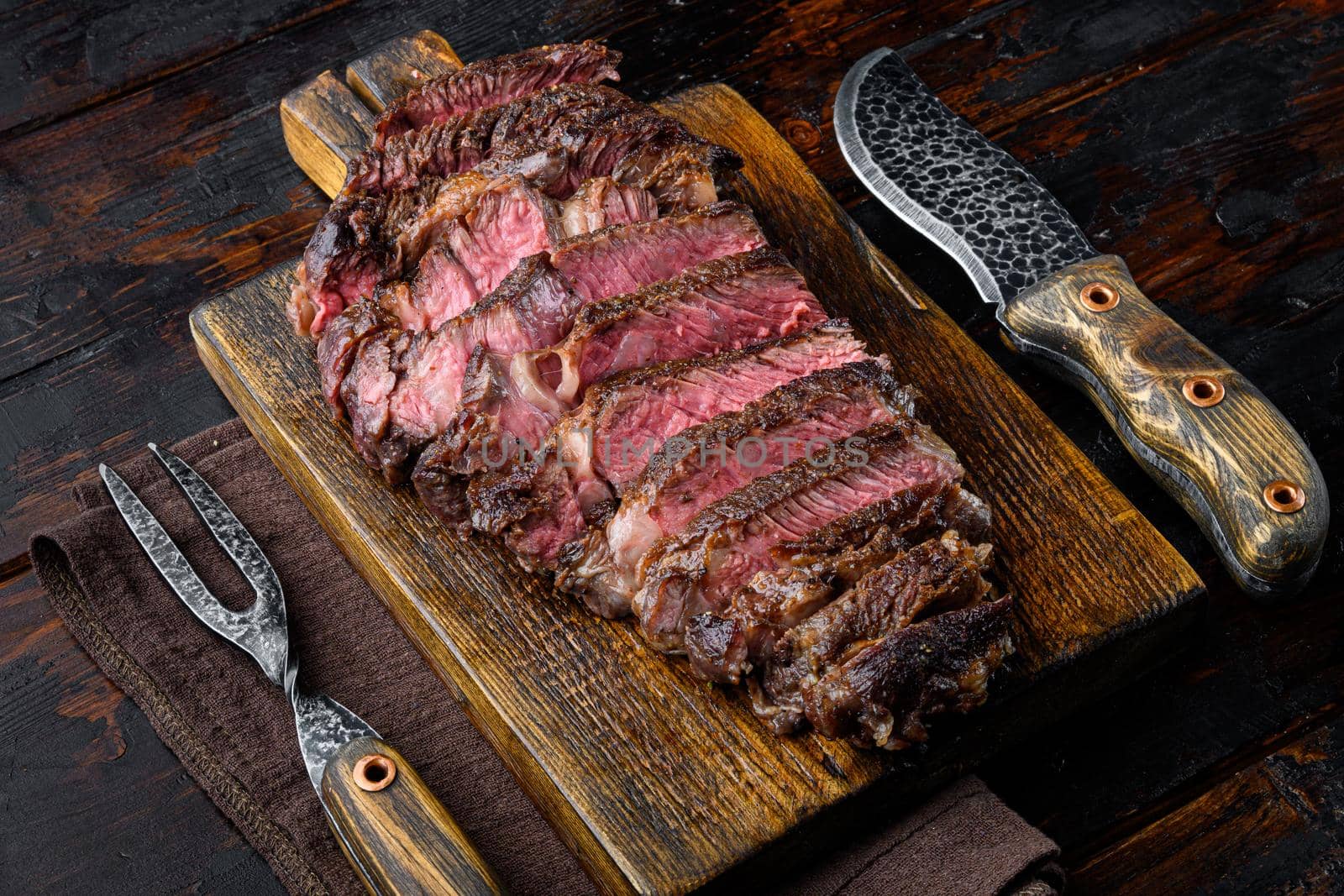 Juicy medium beef Rib Eye steak slices set, on wooden serving board, with meat knife and fork, on old dark wooden table background
