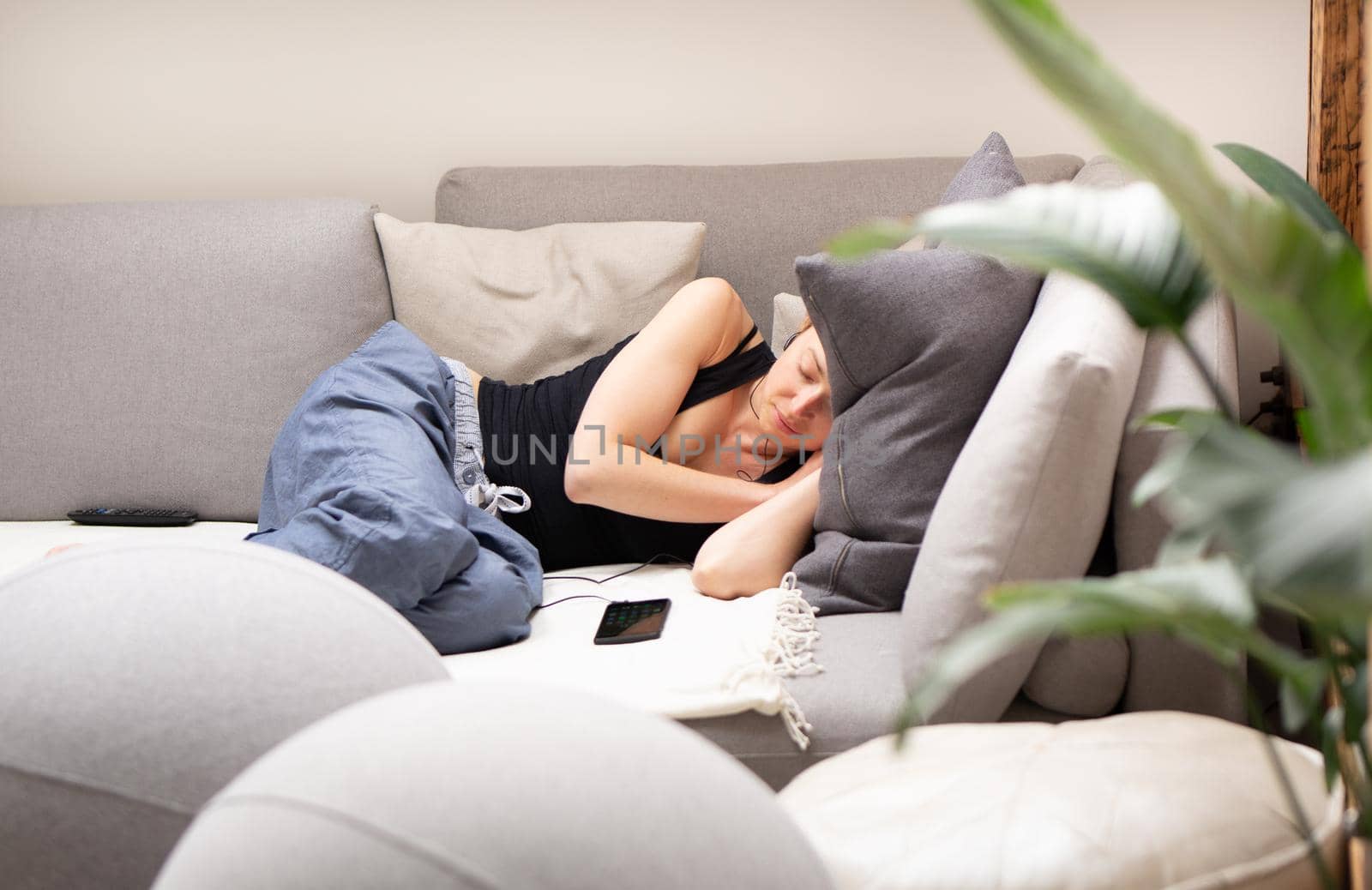 Tired young woman taking a nap at home lying on a comfortable sofa with big cushions in living room with a mobile phone lying next to her. Healthy lifestyle concept.