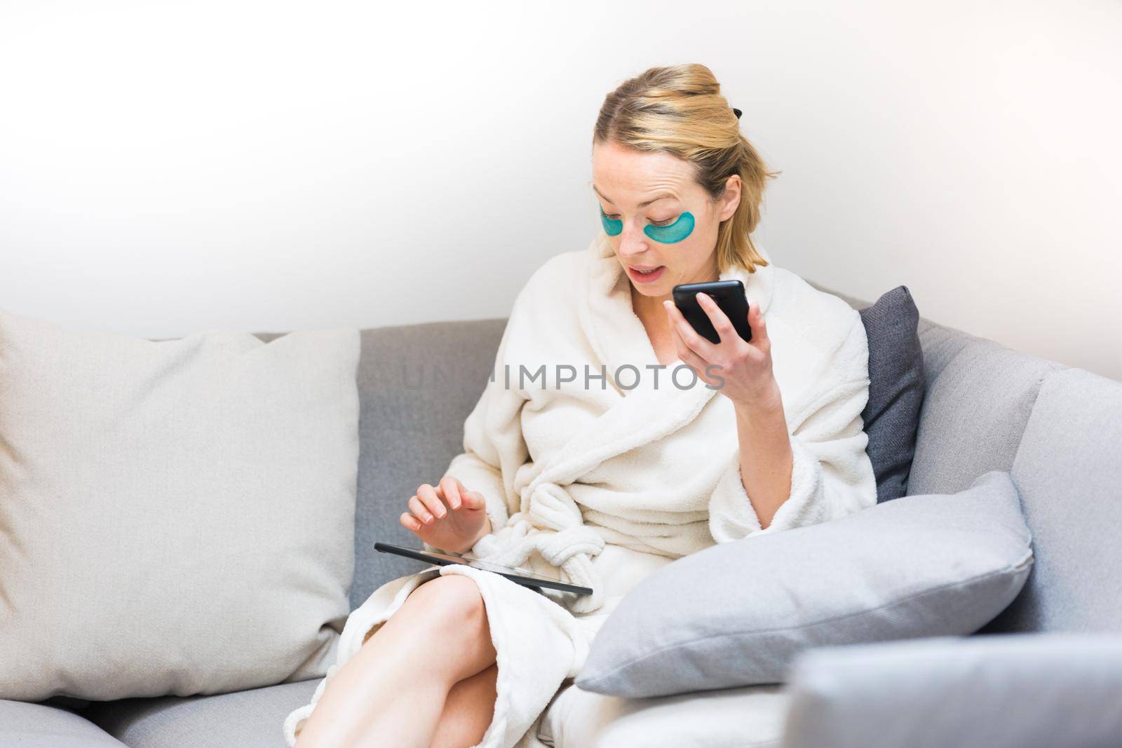 Young female entrepreneur wearing beauty face mask and cosy warm bathrobe in the morning, working remotly from her living room sofa. Remote work from home concept