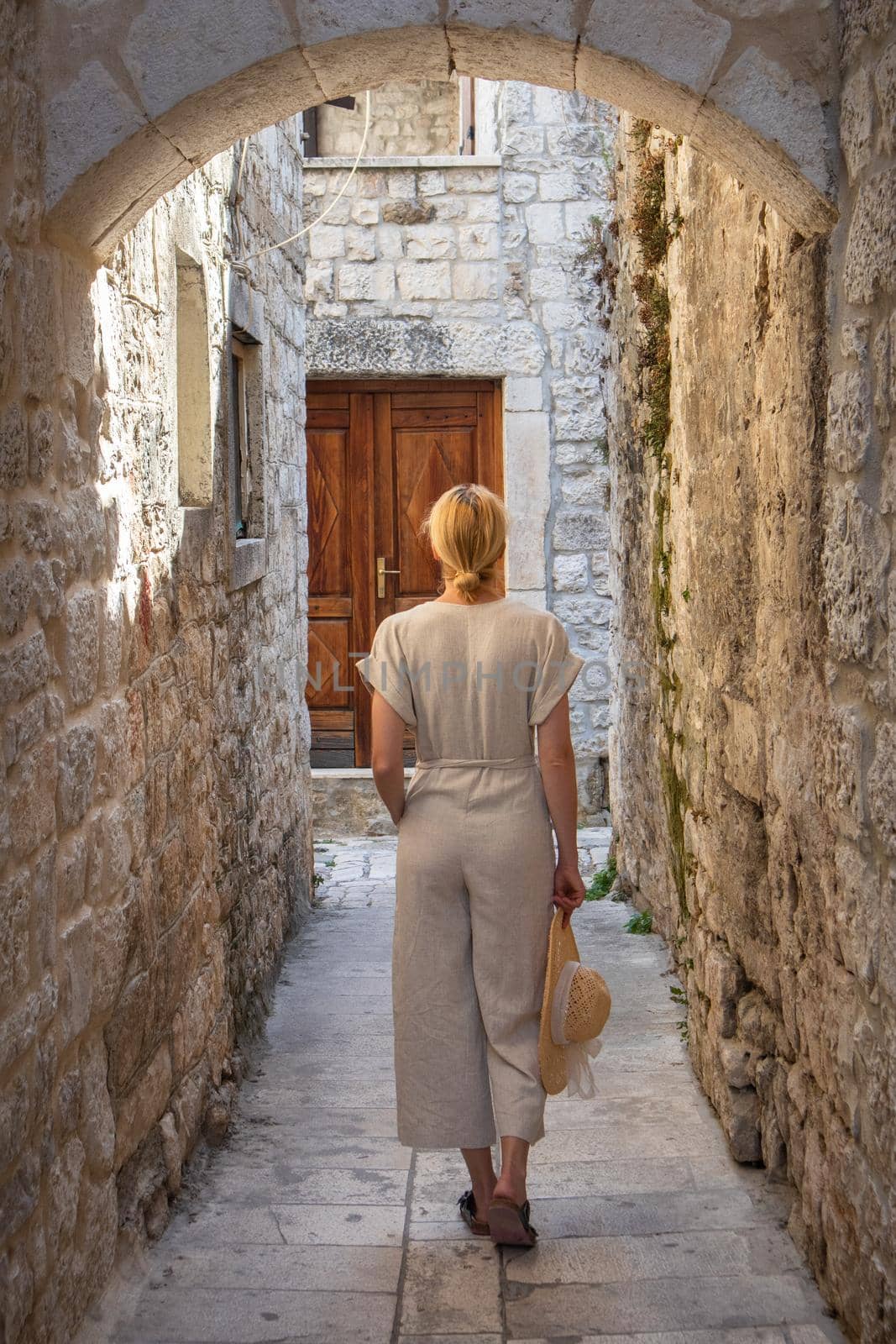 Rear view of beautiful blonde young female traveler wearing straw sun hat sightseeing and enjoying summer vacation in an old traditional costal town at Adriatic cost, Croatia.