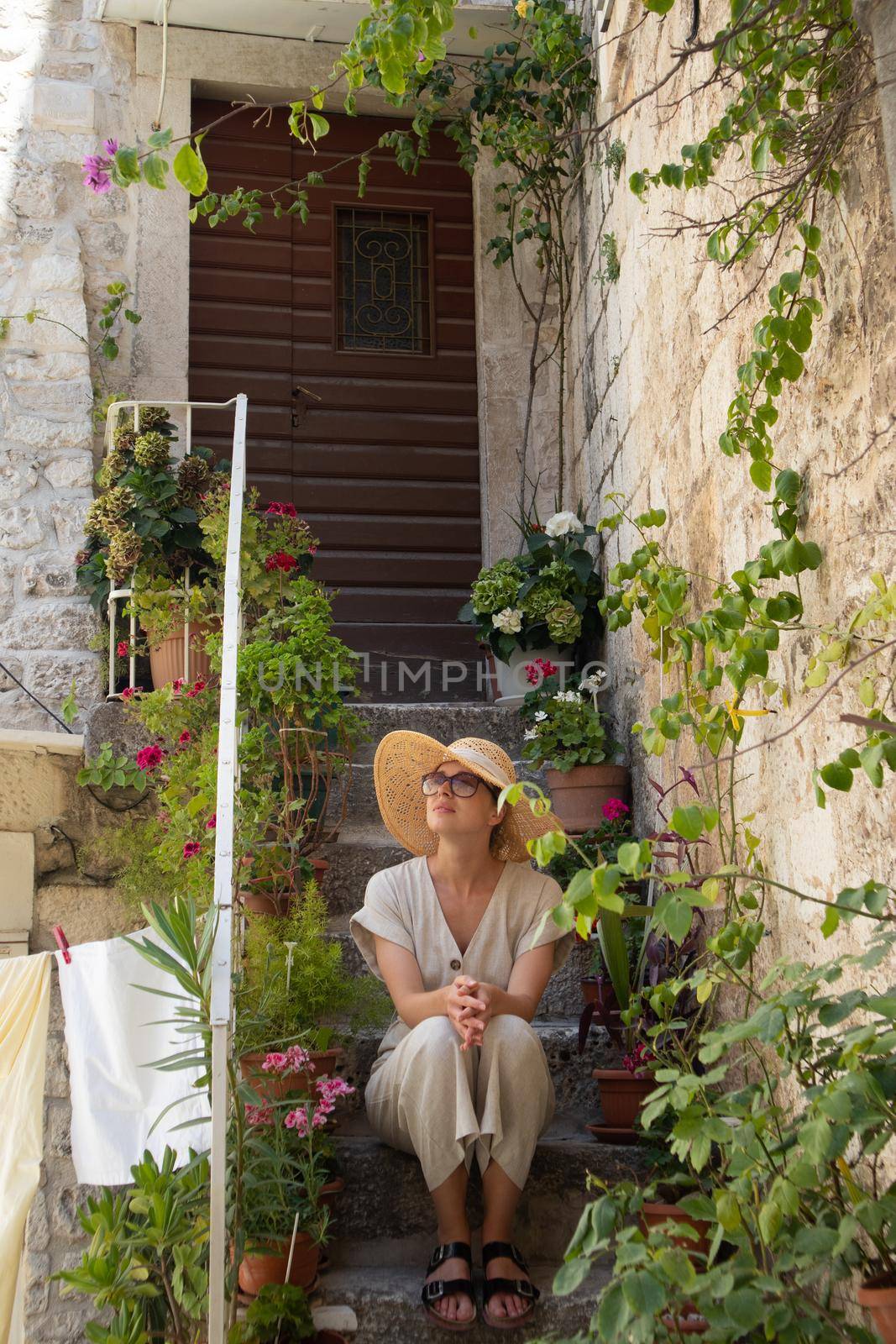 Beautiful female tourist wearing big straw sun hat and sundress sitting and relaxing on old stone house stairs during summer travel on Mediterranean cost on hot summer day