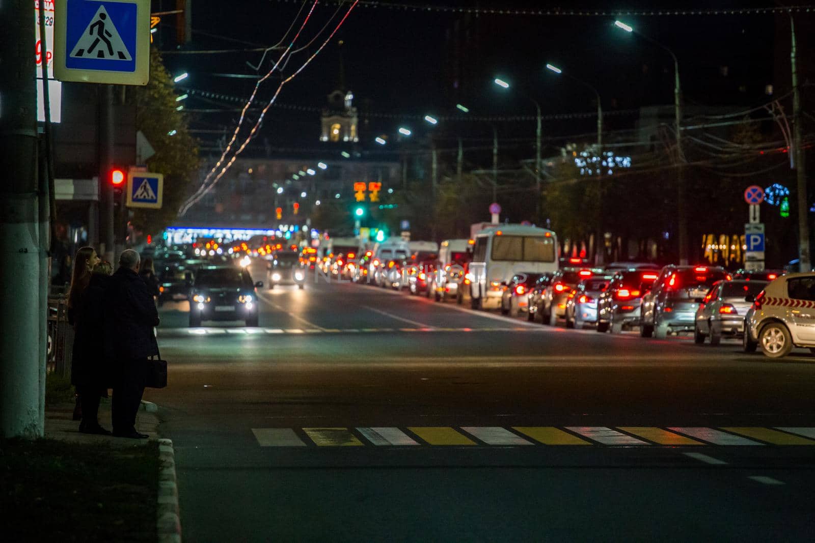 TULA, RUSSIA - OCTOBER 16, 2018: citizens waiting signal for crossing central prospect at night in light of traffic with blurry background jam