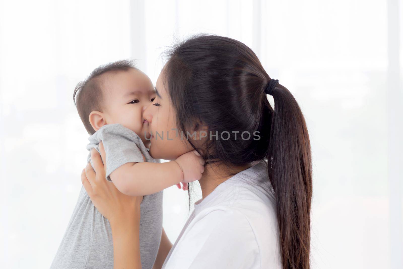 Young asian mother holding little baby girl and kiss together in the bedroom at home, mom carry child and care, woman and kid with carefree, toddler and parent, emotion and expression, family concept.