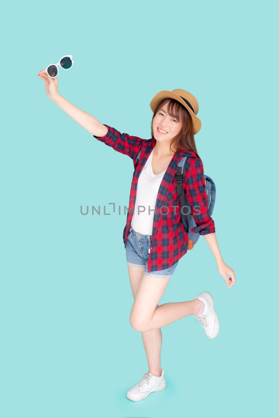Beautiful portrait young asian woman wear hat and holding sunglasses smile expression confident enjoy summer holiday isolated blue background, model girl fashion having backpack, travel concept.