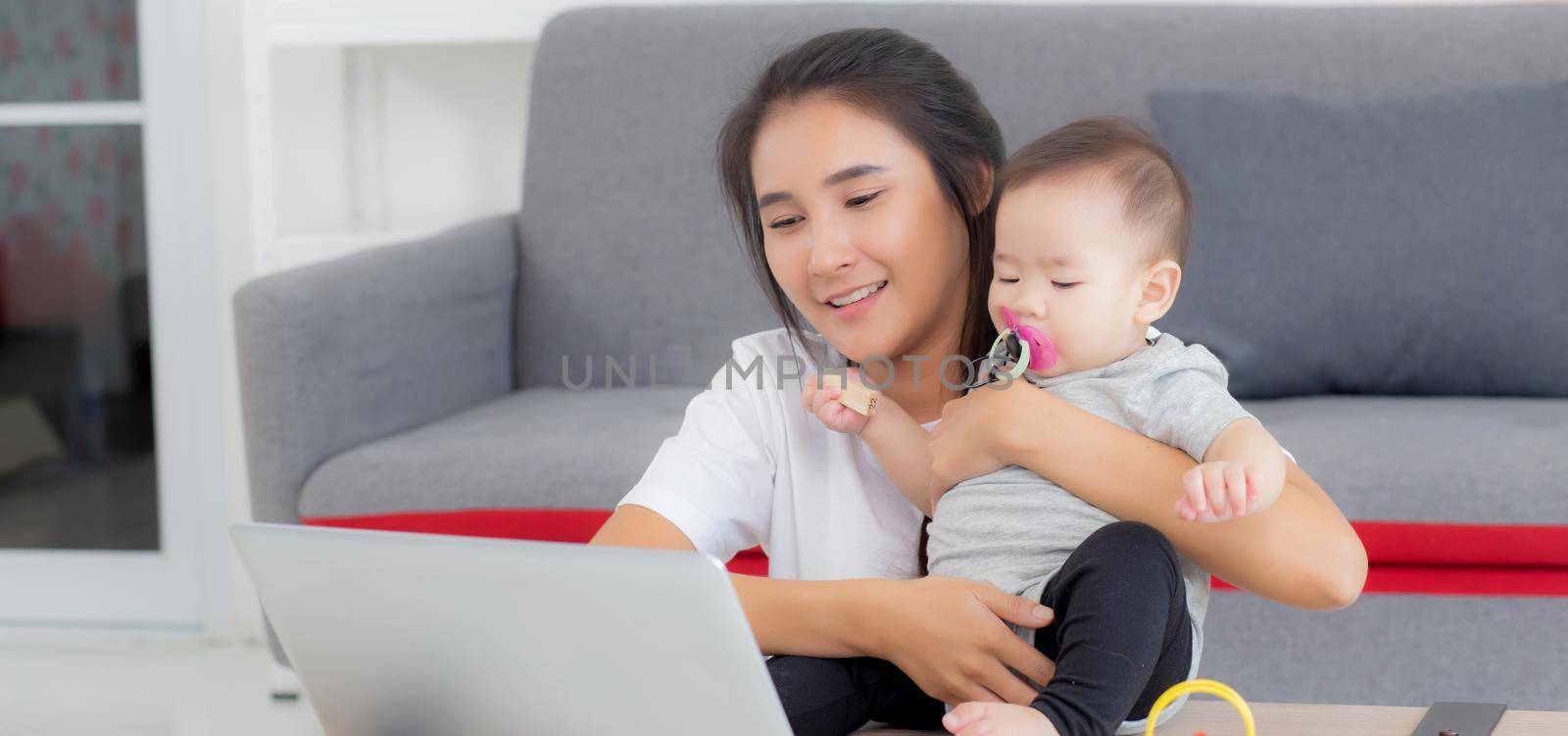 Young asian mother working on laptop computer while care daughter girl at home, mom and baby girl sitting using notebook video call, parent and bonding, indoors, family and business concept. by nnudoo