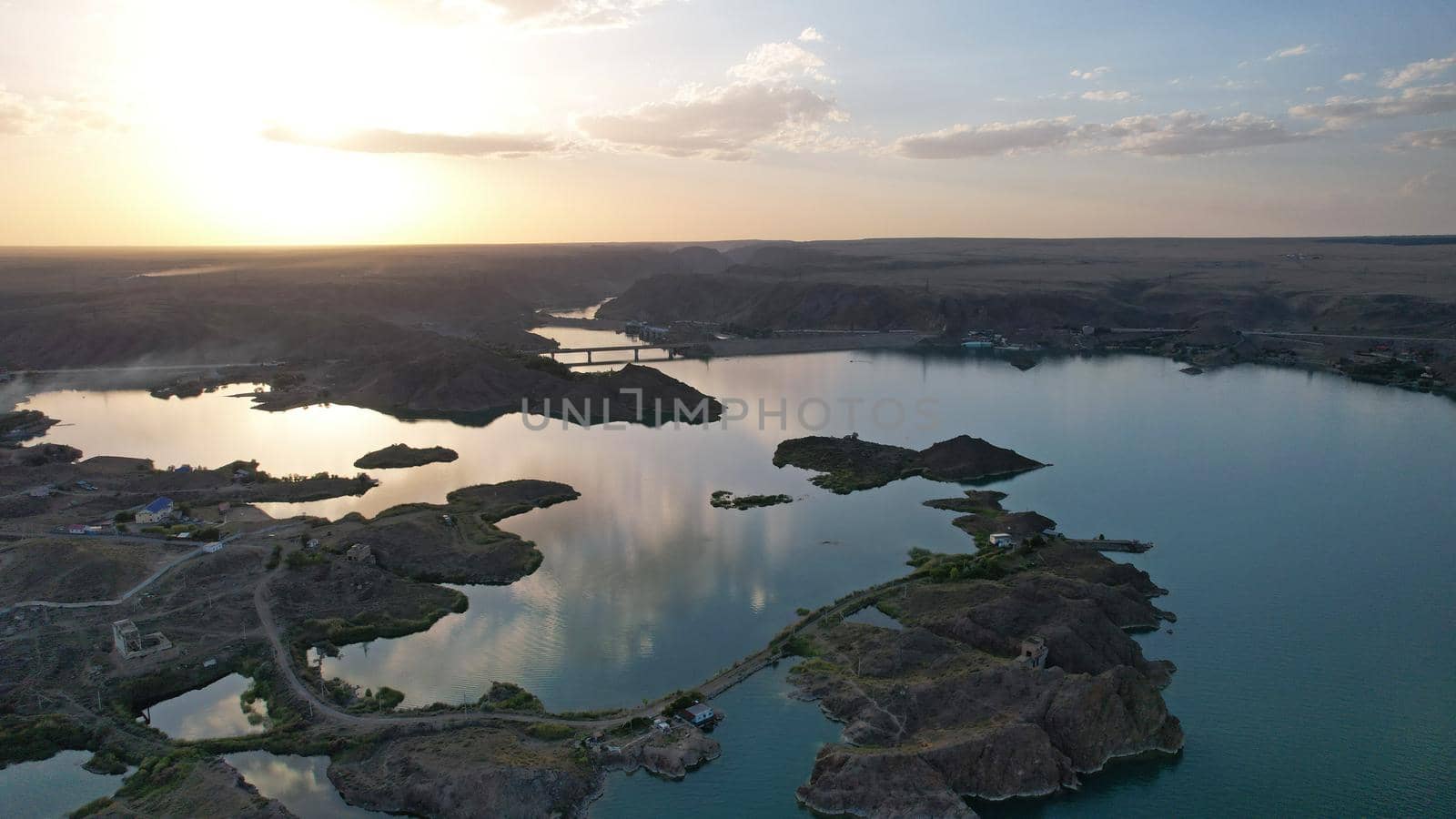 A speedboat sails along the strait at sunset. Top view from the drone. A long plume on the water. You can see the bridge and the rocky shore. The water reflects clouds and the sky. Kapchagai reservoir