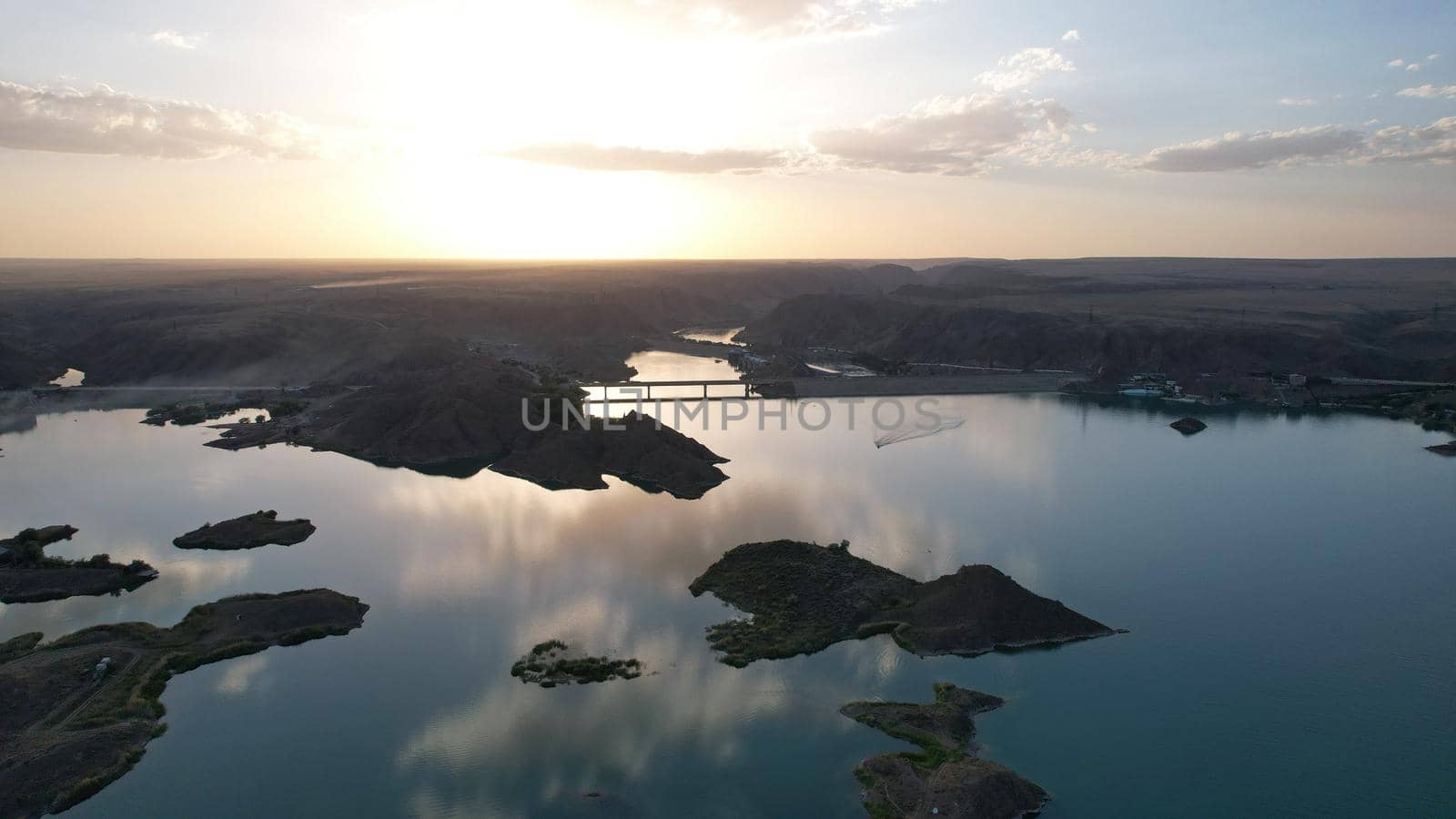 A speedboat sails along the strait at sunset. Top view from the drone. A long plume on the water. You can see the bridge and the rocky shore. The water reflects clouds and the sky. Kapchagai reservoir