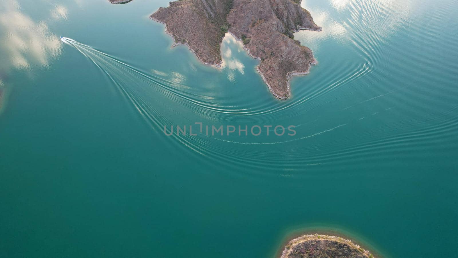 A speedboat sails along the strait at sunset. Top view from the drone. A long plume on the water. You can see the bridge and the rocky shore. The water reflects clouds and the sky. Kapchagai reservoir