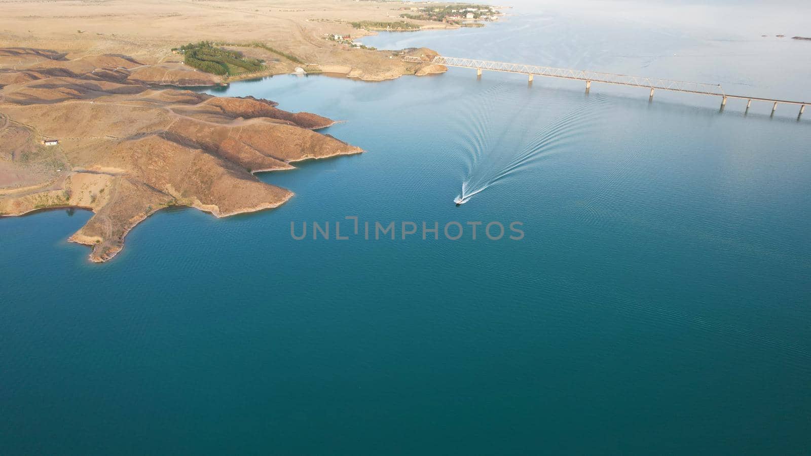 Speedboats sail along the strait. Top view from the drone. Plume on the water. You can see the bridge over the lake, the rocky shore. The water reflects the clouds and the sky. Kapchagai reservoir.