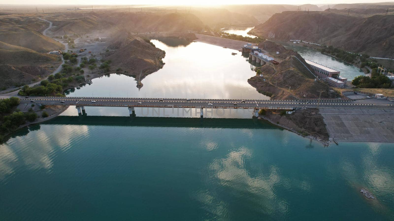 Sunset with a view of the causeway and the bridge over the strait. Cars drive on the bridge. The water reflects the sun, sky and clouds. There are houses on the rocky shore. Kapchagai reservoir.