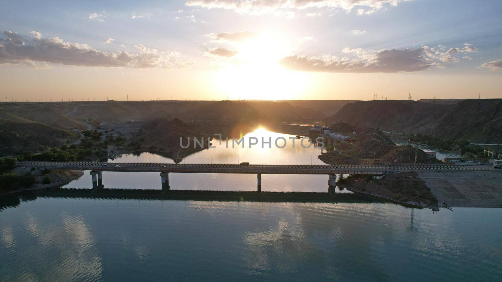 Sunset with a view of the causeway and the bridge over the strait. Cars drive on the bridge. The water reflects the sun, sky and clouds. There are houses on the rocky shore. Kapchagai reservoir.