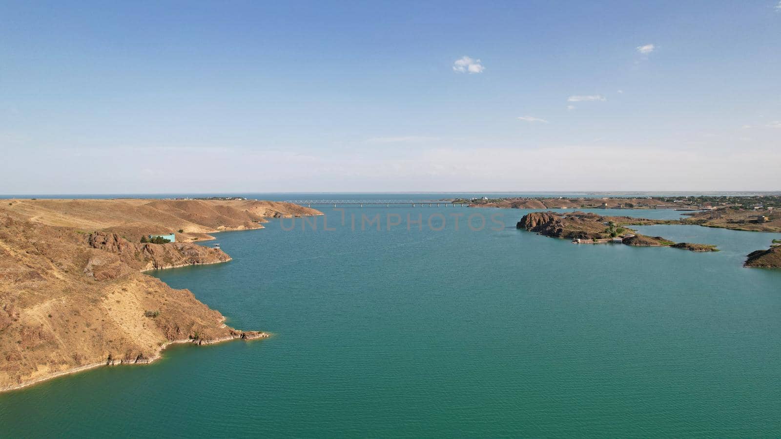 Flying over a lake with islands and rocks. Green water reflects the rays of the sun. Trees and bushes grow on the rocky ground, and houses are built. White clouds. Kapchagai Reservoir, Kazakhstan.