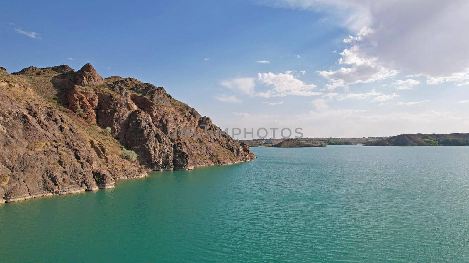 Flying over a lake with islands and rocks. Green water reflects the rays of the sun. Trees and bushes grow on the rocky ground, and houses are built. White clouds. Kapchagai Reservoir, Kazakhstan.