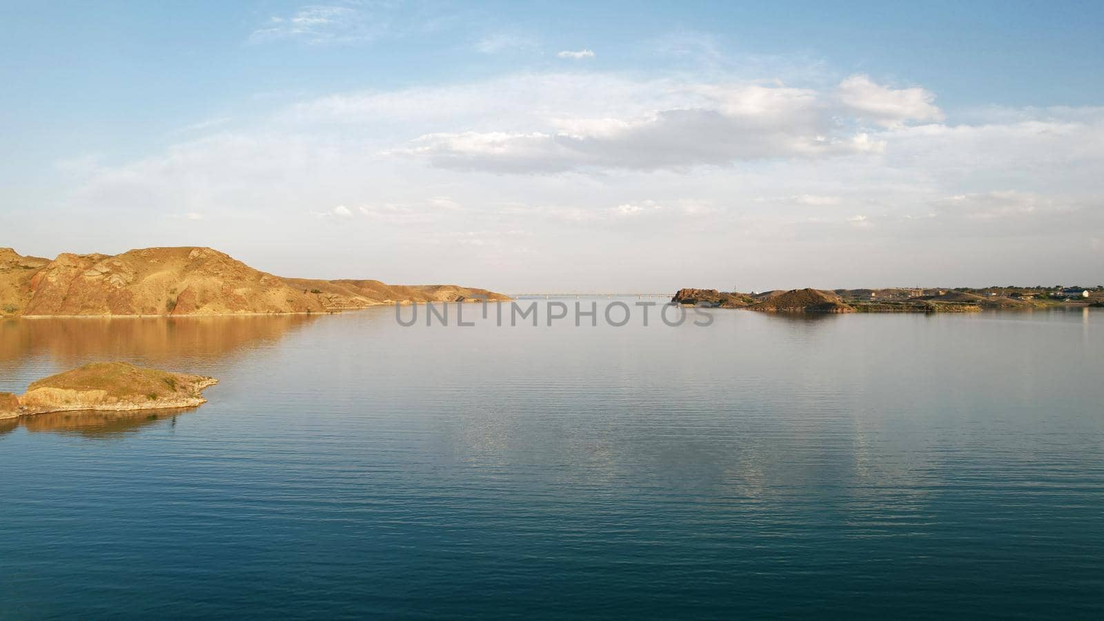 Flying over a lake with islands and rocks. Green water reflects the rays of the sun. Trees and bushes grow on the rocky ground, and houses are built. White clouds. Kapchagai Reservoir, Kazakhstan.