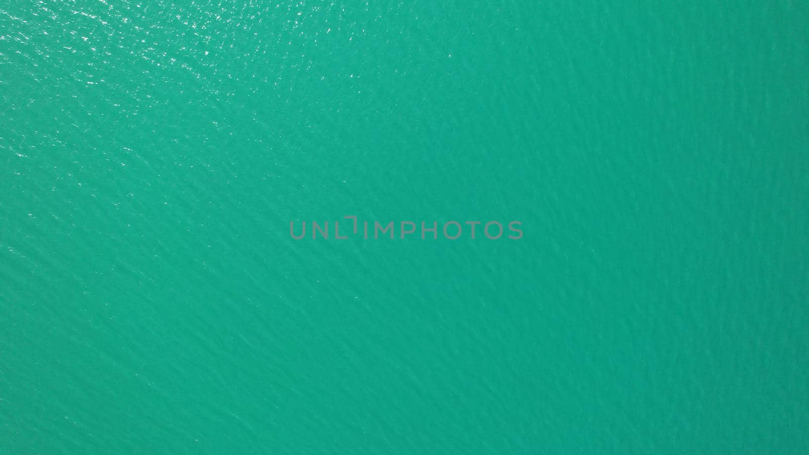 Top view of green water and small waves. The background of the water shimmers in the rays of the sun with highlights. White foam is visible. The gradient of the lake and the beach. Rocks on the bottom