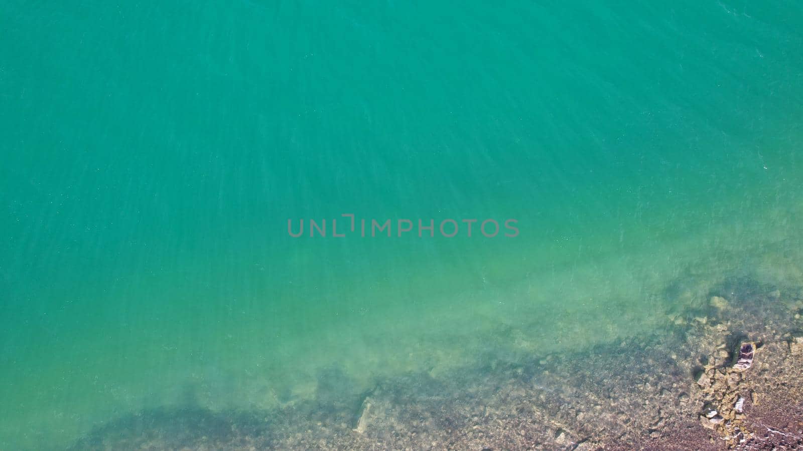 Top view of green water and small waves. The background of the water shimmers in the rays of the sun with highlights. White foam is visible. The gradient of the lake and the beach. Rocks on the bottom