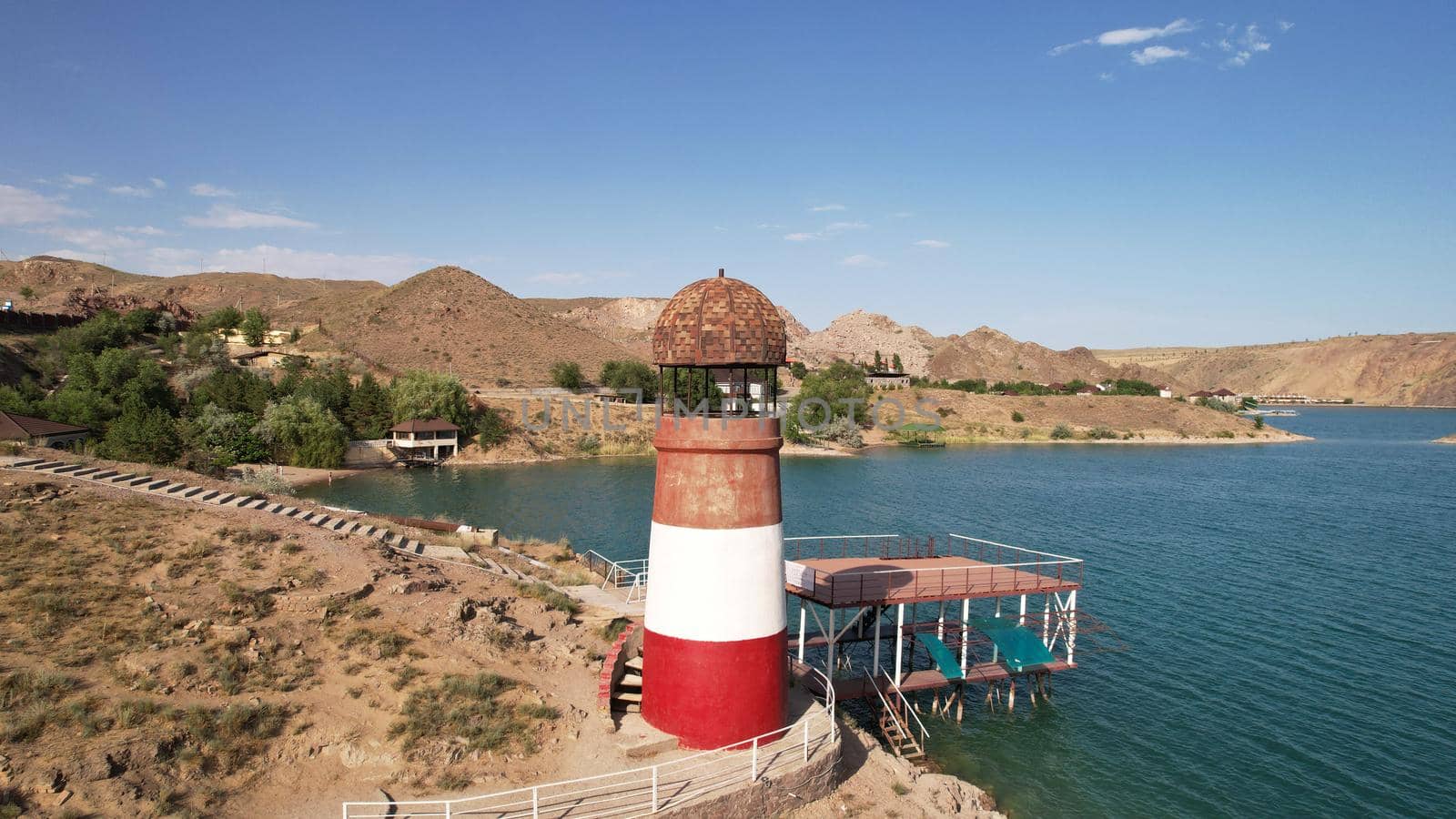 White and red lighthouse on the shore of the lake. The green water of the reservoir reflects the sky and clouds. Rocky beach. In some places, trees and bushes grow. Shtul. Kapchagai, Kazakhstan.