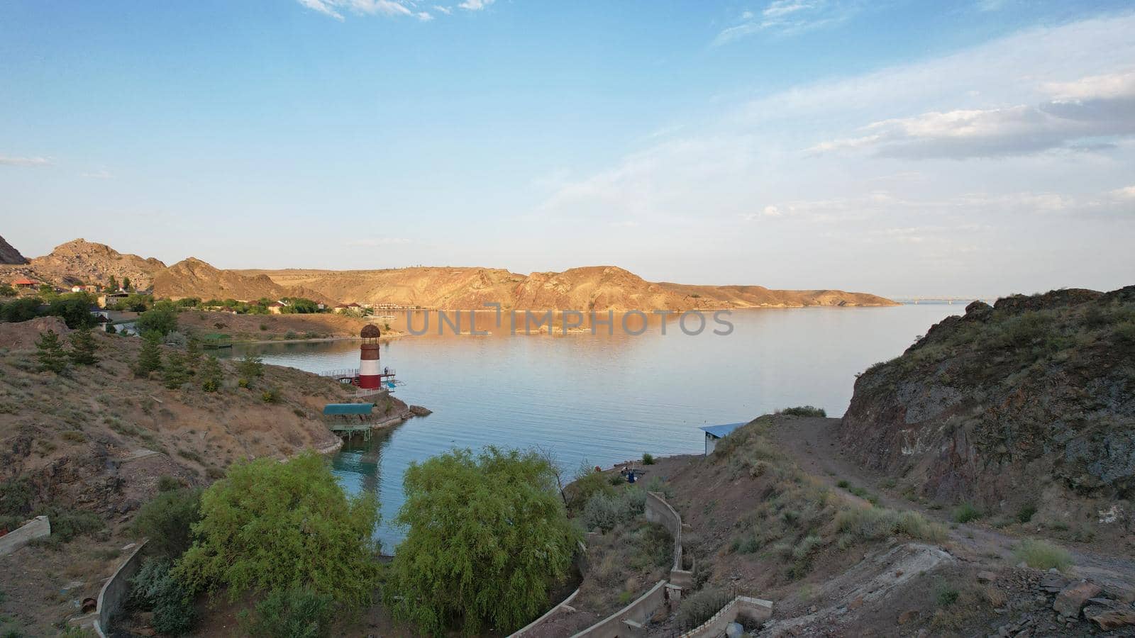 White and red lighthouse on the shore of the lake. The green water of the reservoir reflects the sky and clouds. Rocky beach. In some places, trees and bushes grow. Shtul. Kapchagai, Kazakhstan.