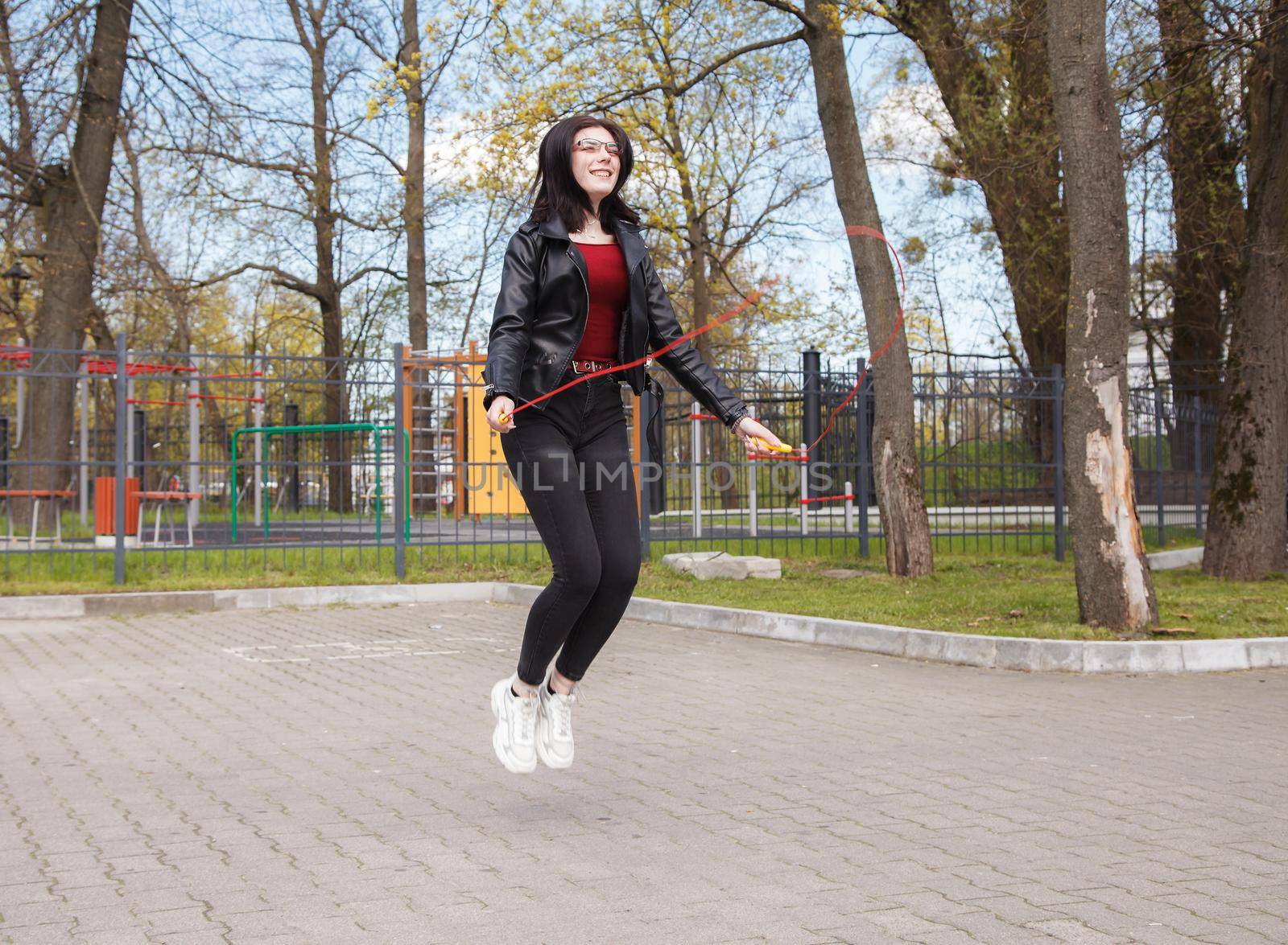 young brunette girl in black jacket and jeans jumping rope outdoor on sunny spring day