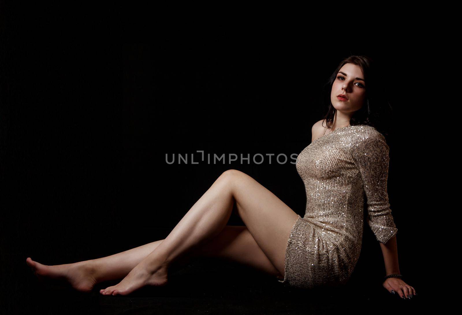 young beautiful brunette girl in bright dress posing sitting on floor in the studio on black background