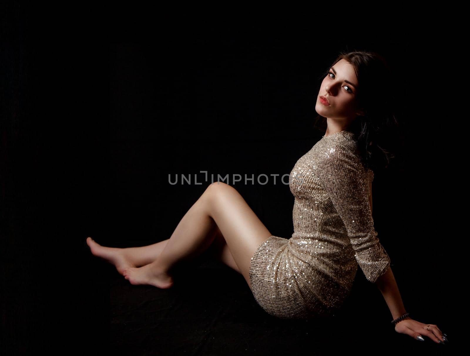 young beautiful brunette girl in bright dress posing sitting on floor in the studio on black background