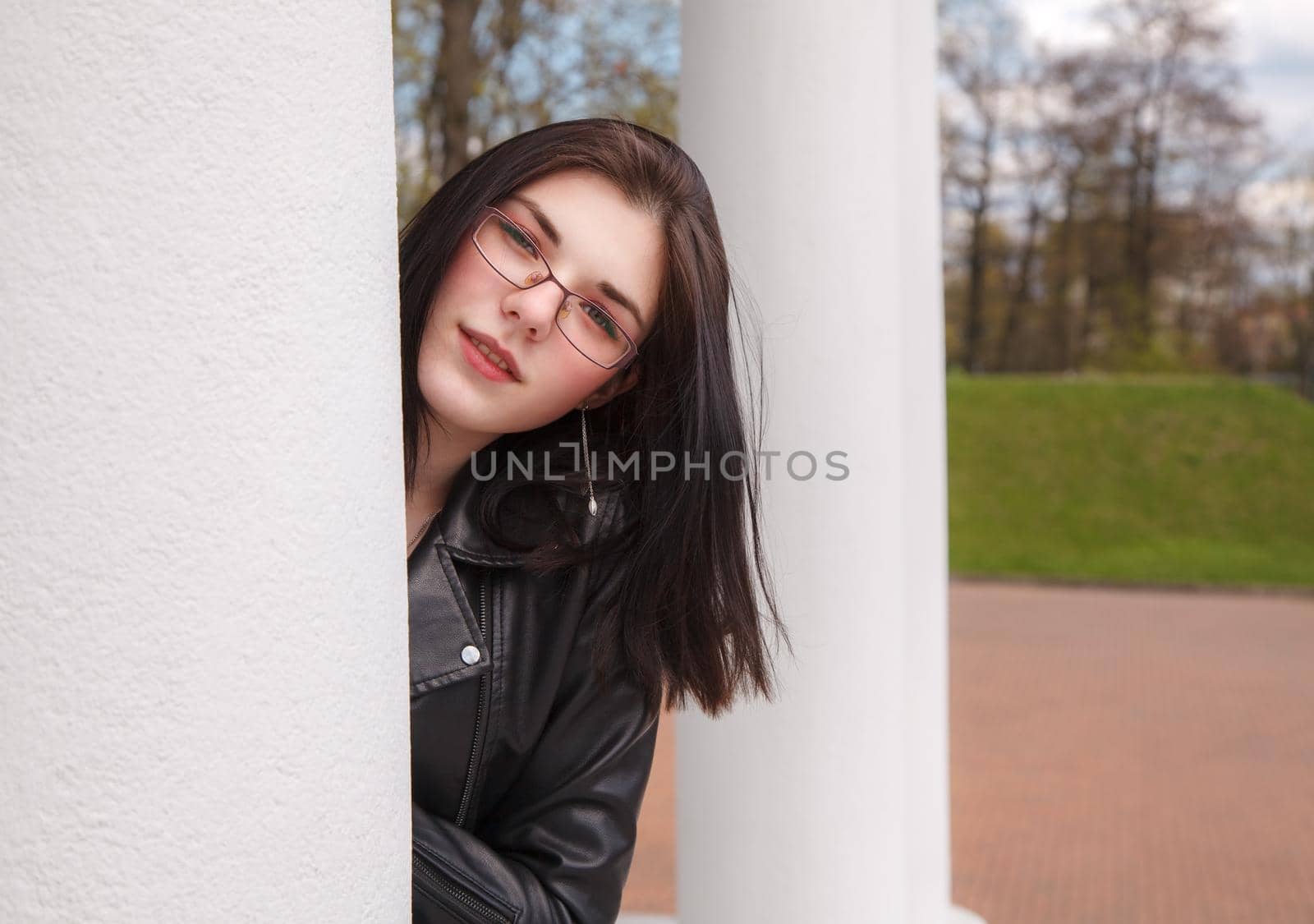 young brunette beautiful in black leather jacket girl peeking out from behind pillar in city park. outdoor sunny spring day
