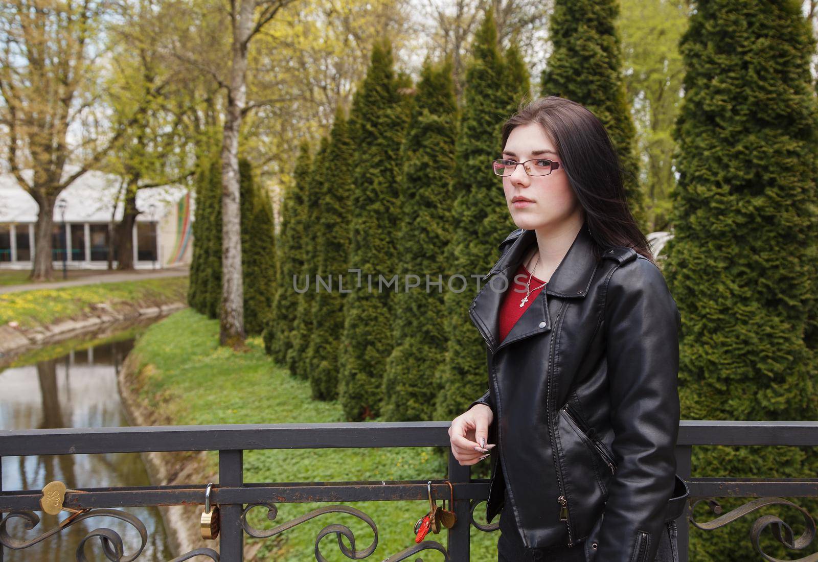 young beautiful brunette girl in black jacket and jeans stands on the embankment near river in city park on sunny spring day